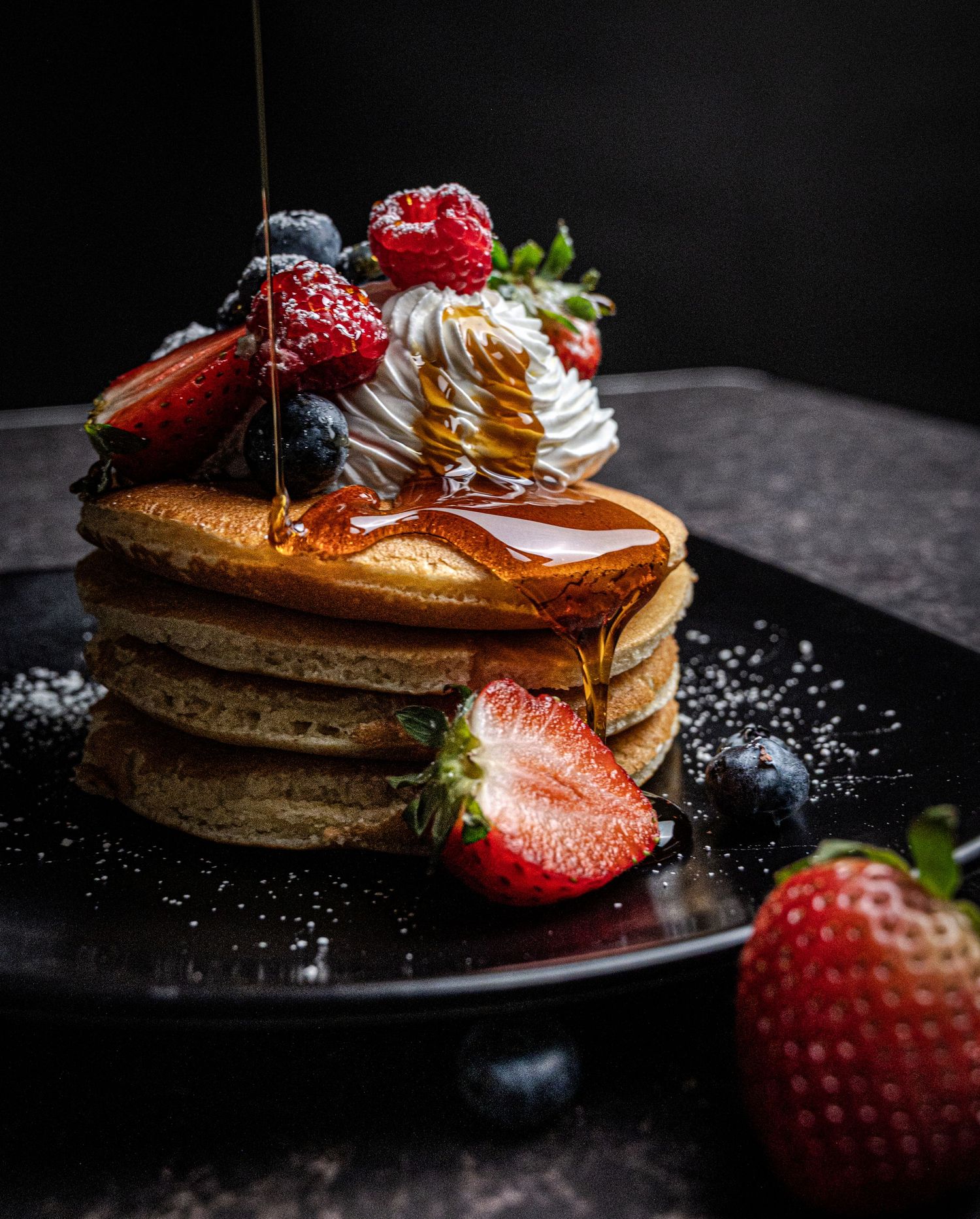 Stack of fluffy pancakes topped with whipped cream, fresh strawberries, blueberries, and drizzled maple syrup on a dark plate.