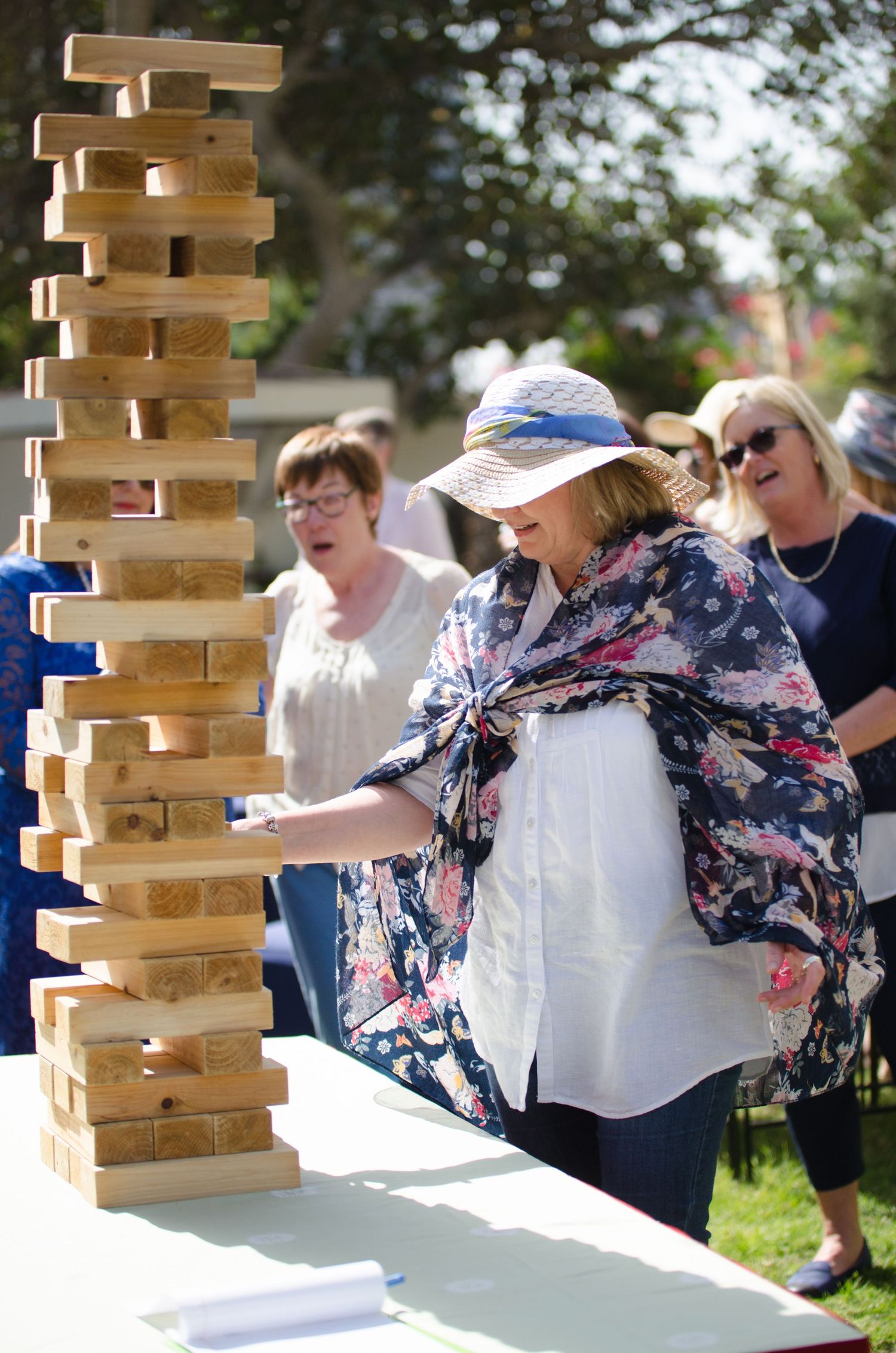 Woman in a floral kimono and sun hat carefully removes a block from a tall wooden giant Jenga tower at an outdoor party as guests watch.