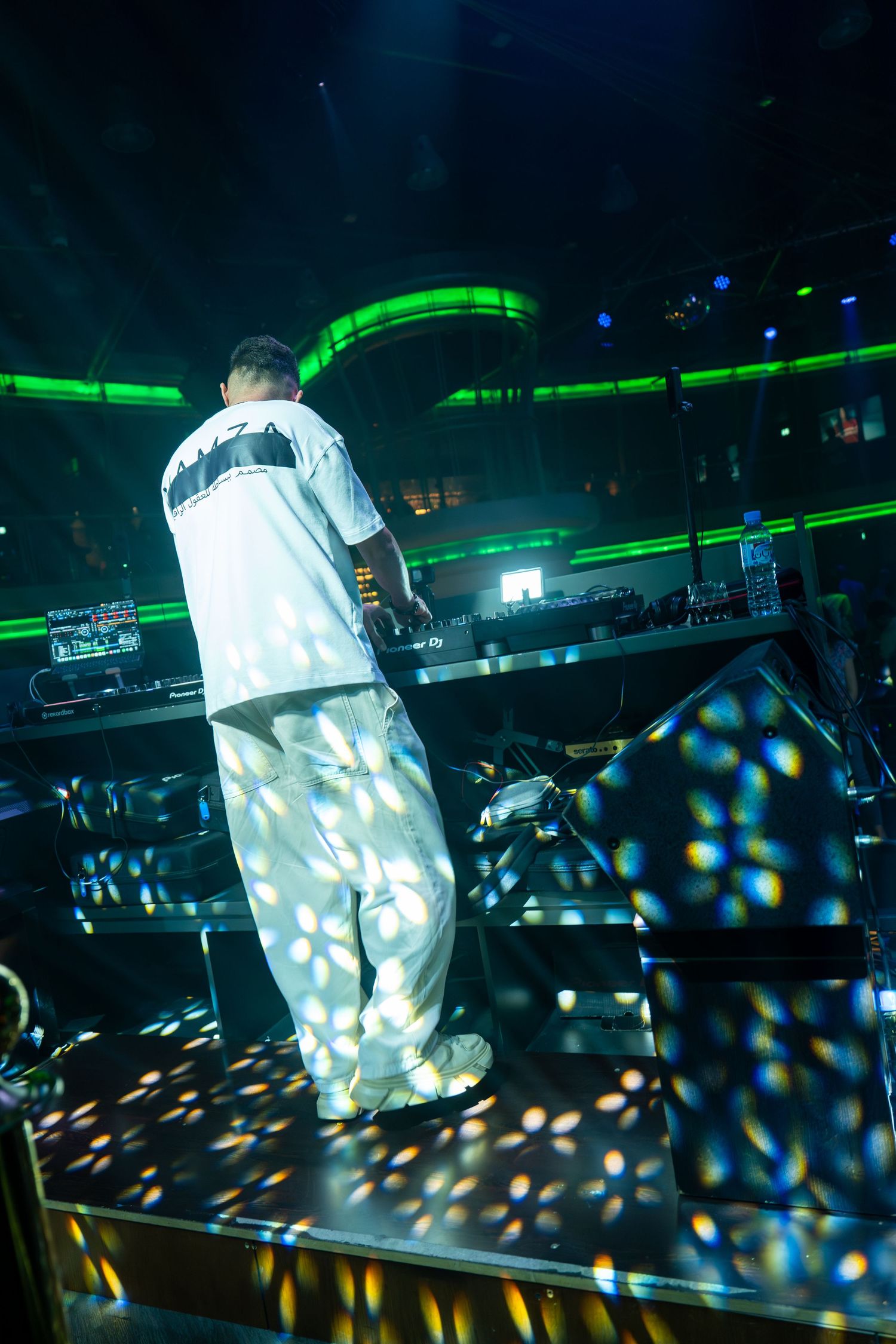 DJ performing on stage in a nightclub with colorful spotlights and green neon lighting effects.