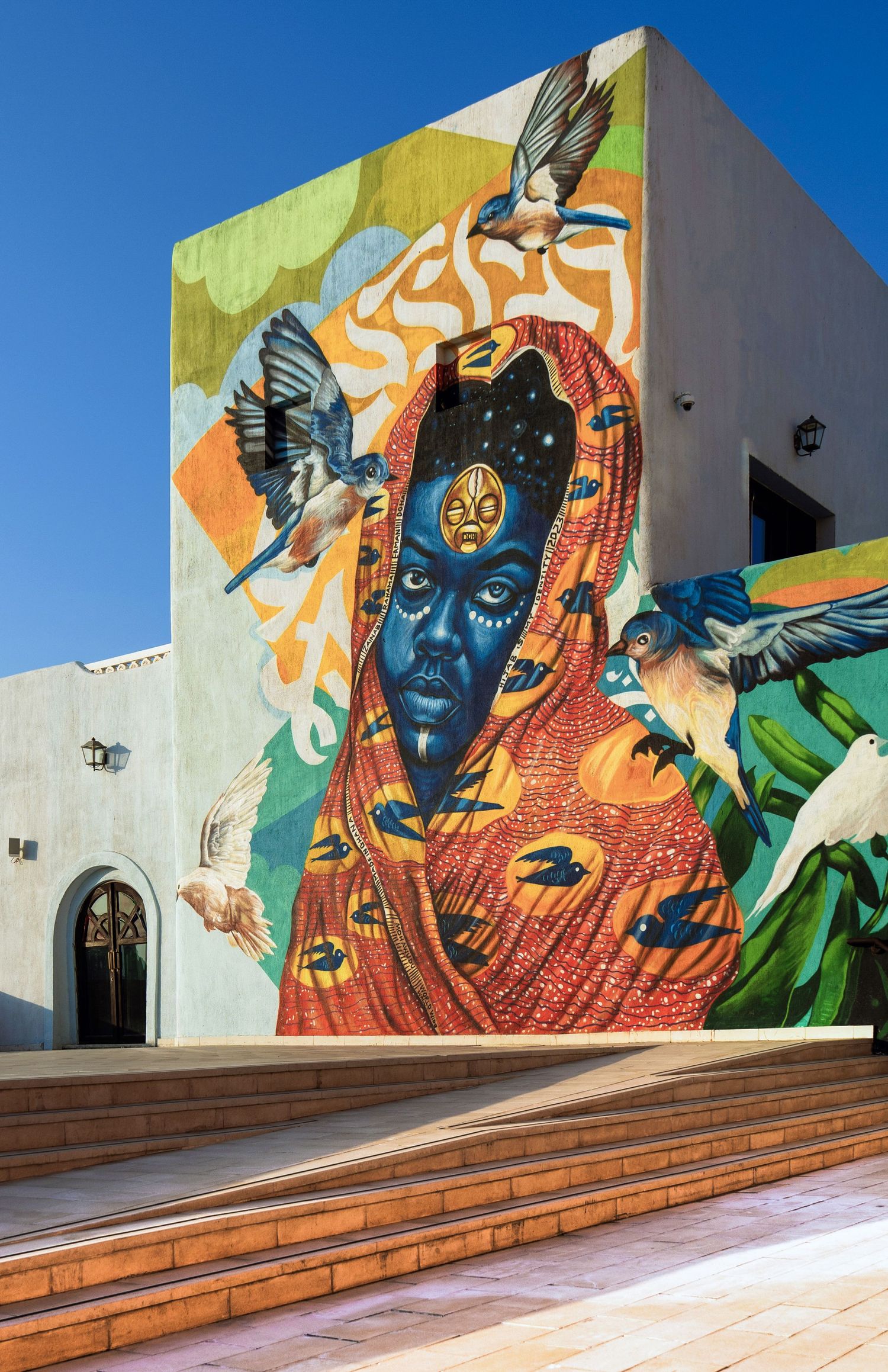 Vibrant street mural of a blue-faced figure in red robes surrounded by birds on a white building under clear blue sky.