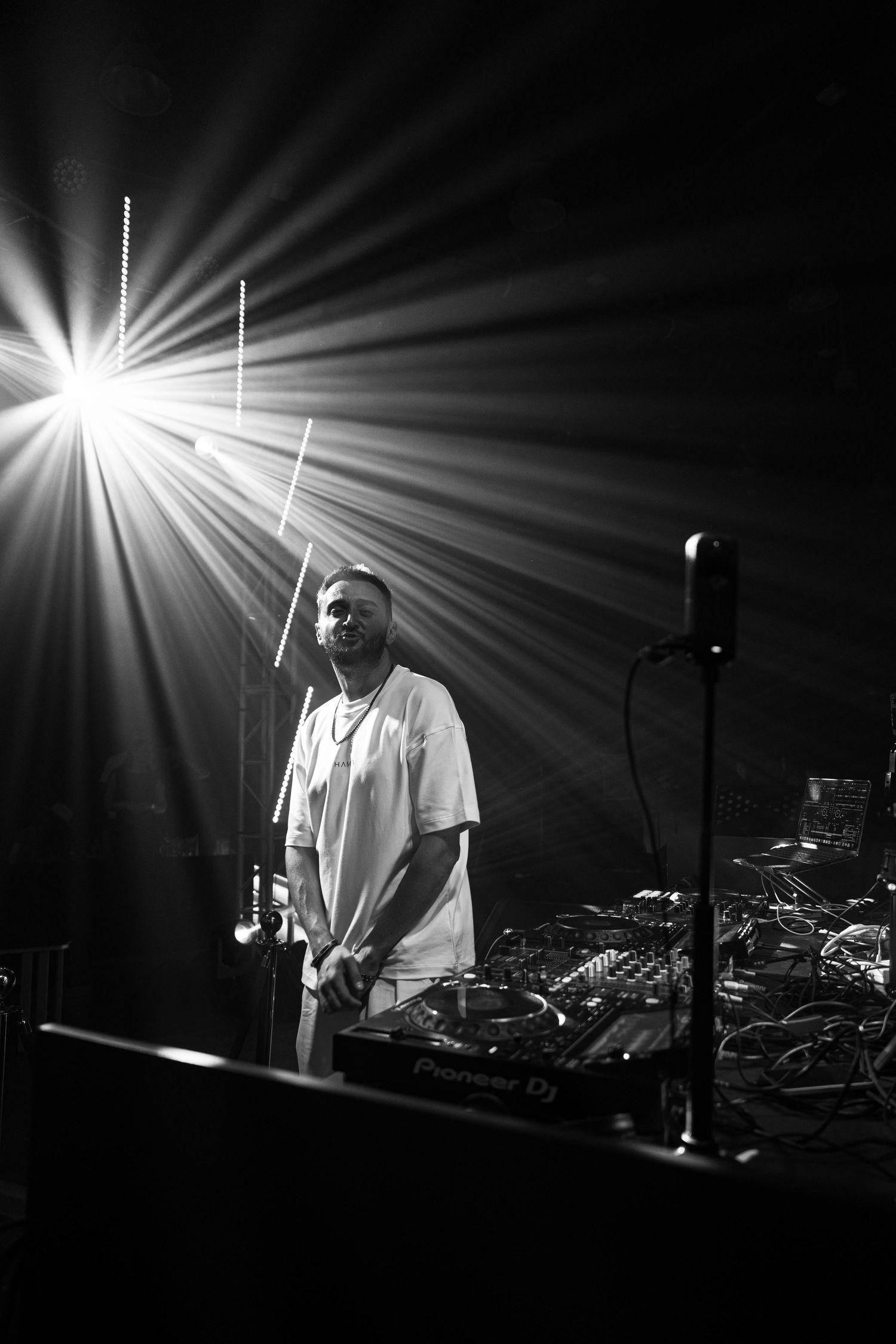 Black and white photo of a DJ standing at Pioneer DJ decks under dramatic stage lighting beams.