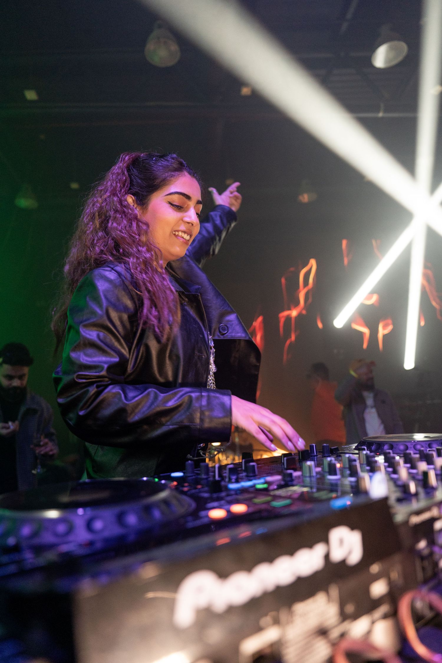 Smiling female DJ with curly hair mixing music on Pioneer DJ equipment at a vibrant nightclub event.
