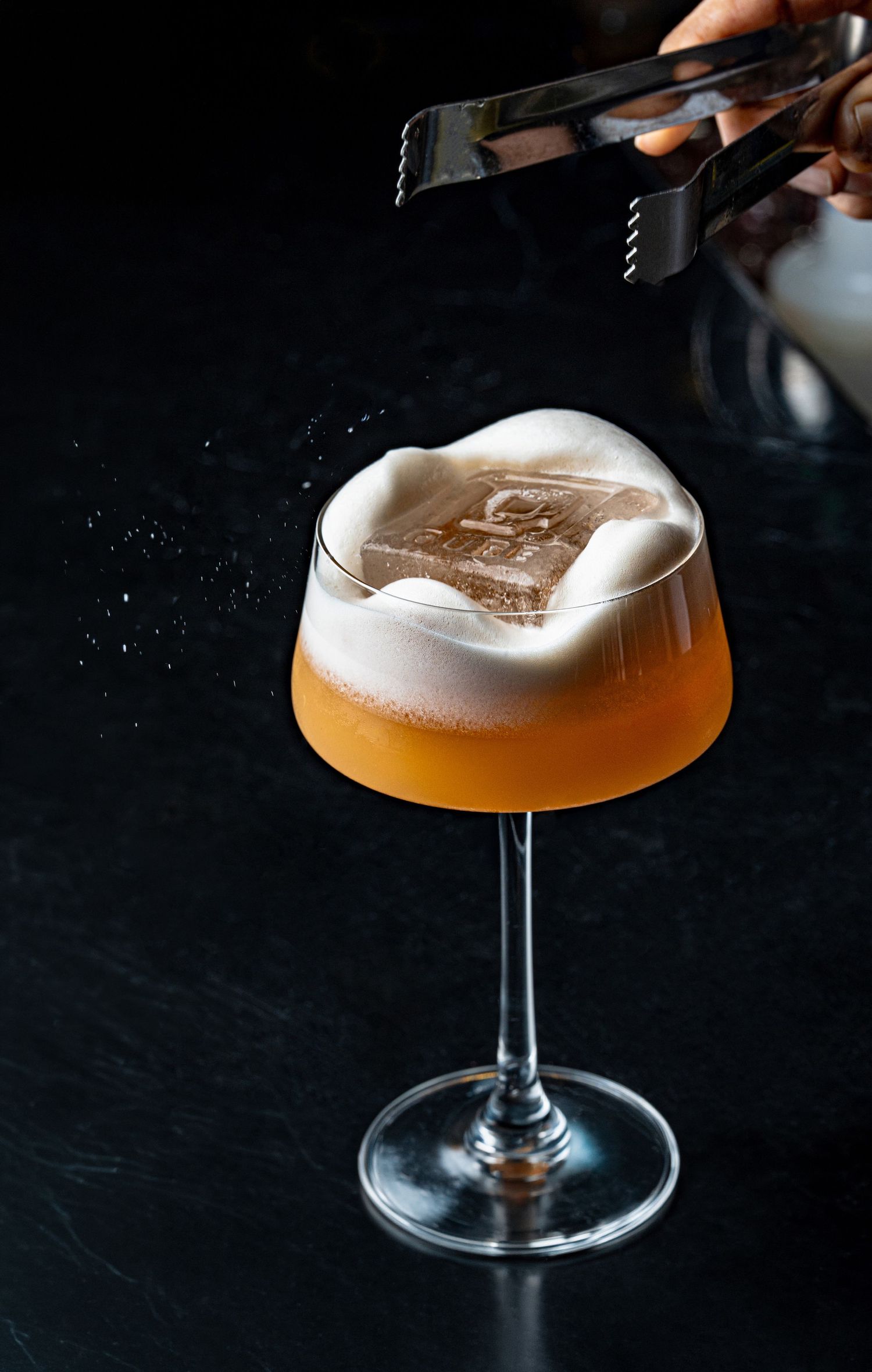 Bartender placing branded ice cube into frothy amber cocktail in elegant coupe glass on dark background.