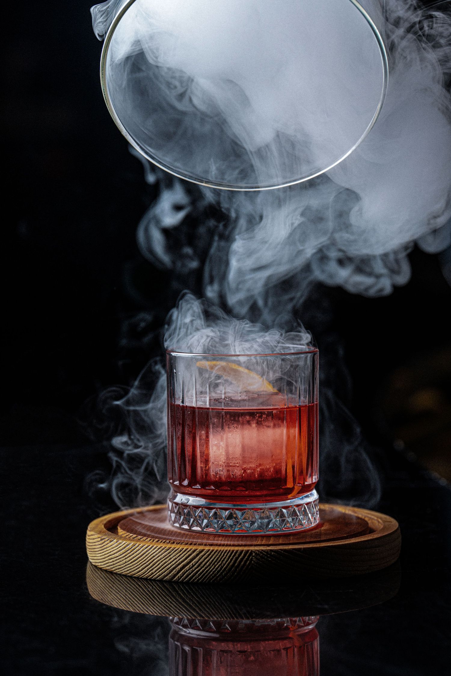 Smoky cocktail in crystal glass on wooden coaster with dramatic smoke swirls on dark background.