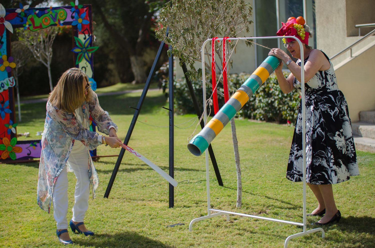 Two women play a festive game on a sunny lawn, one swinging a stick at a colorful striped pole on a white stand near a decorated backdrop.