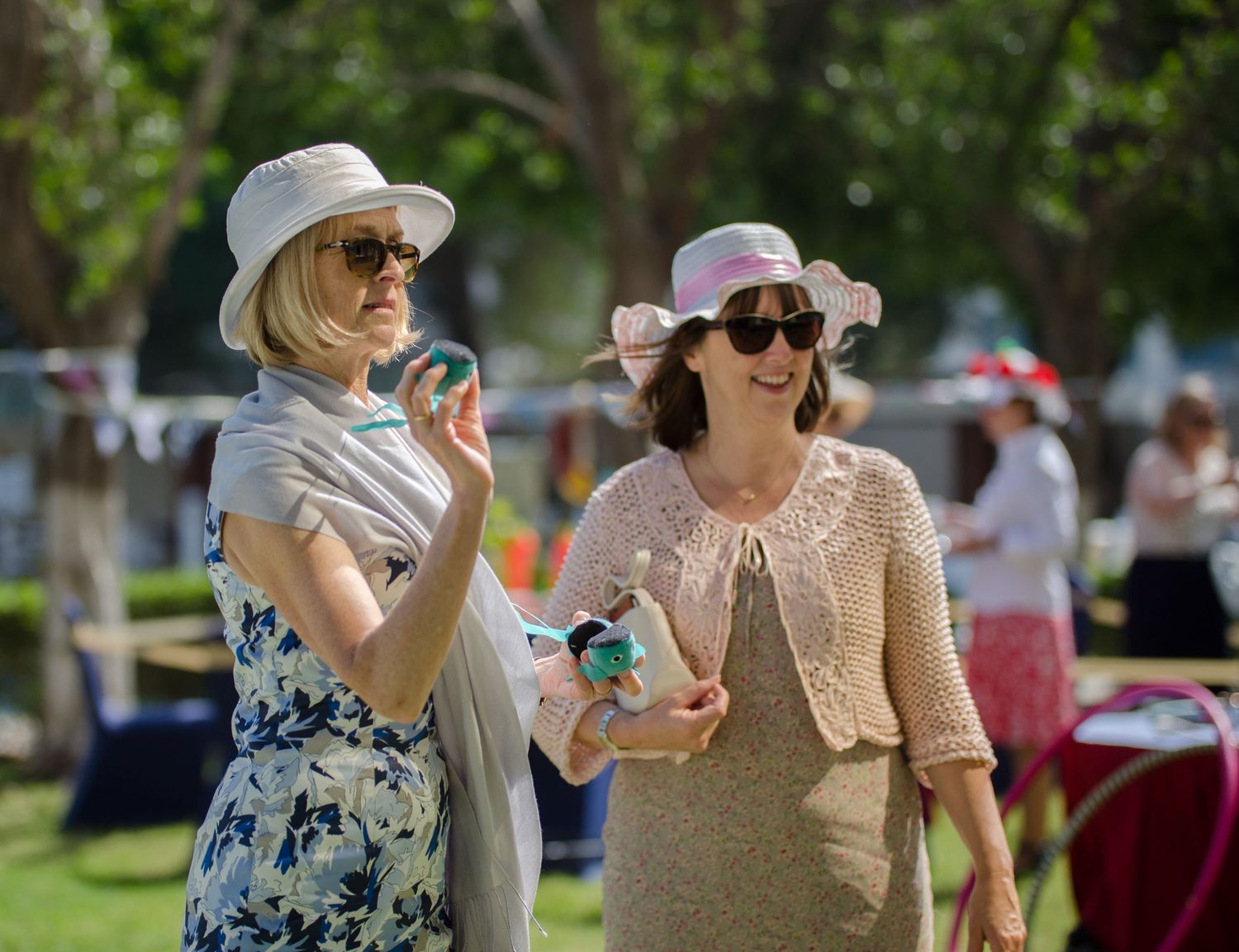 Two women in sun hats and summery outfits chat and hold drinks at a sunny outdoor garden party with tents and trees in the background.