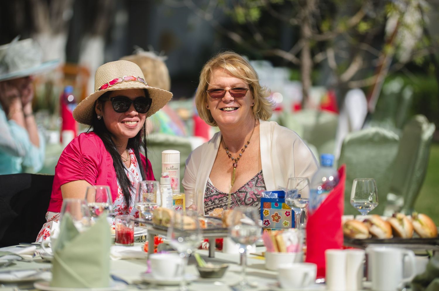 Two smiling women in sunglasses sit at an outdoor garden party table set with glasses, teacups, napkins and pastries.
