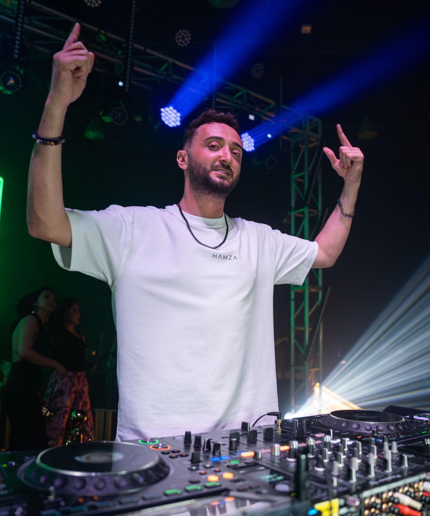 DJ in white tee raises hands at Pioneer DJ decks under blue stage lights at nightclub event.