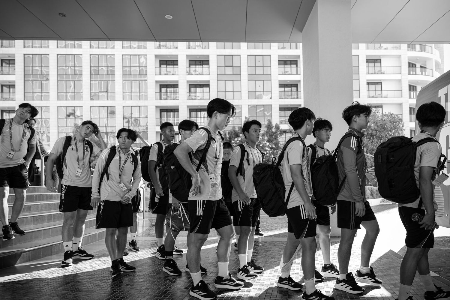 Black and white photo of young athletes with backpacks walking in a line outside a building.