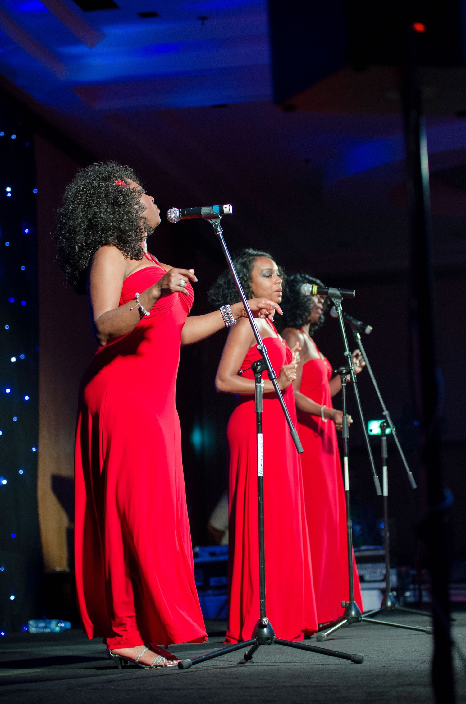 Three women in matching red gowns singing into microphones on a stage at a formal gala event.