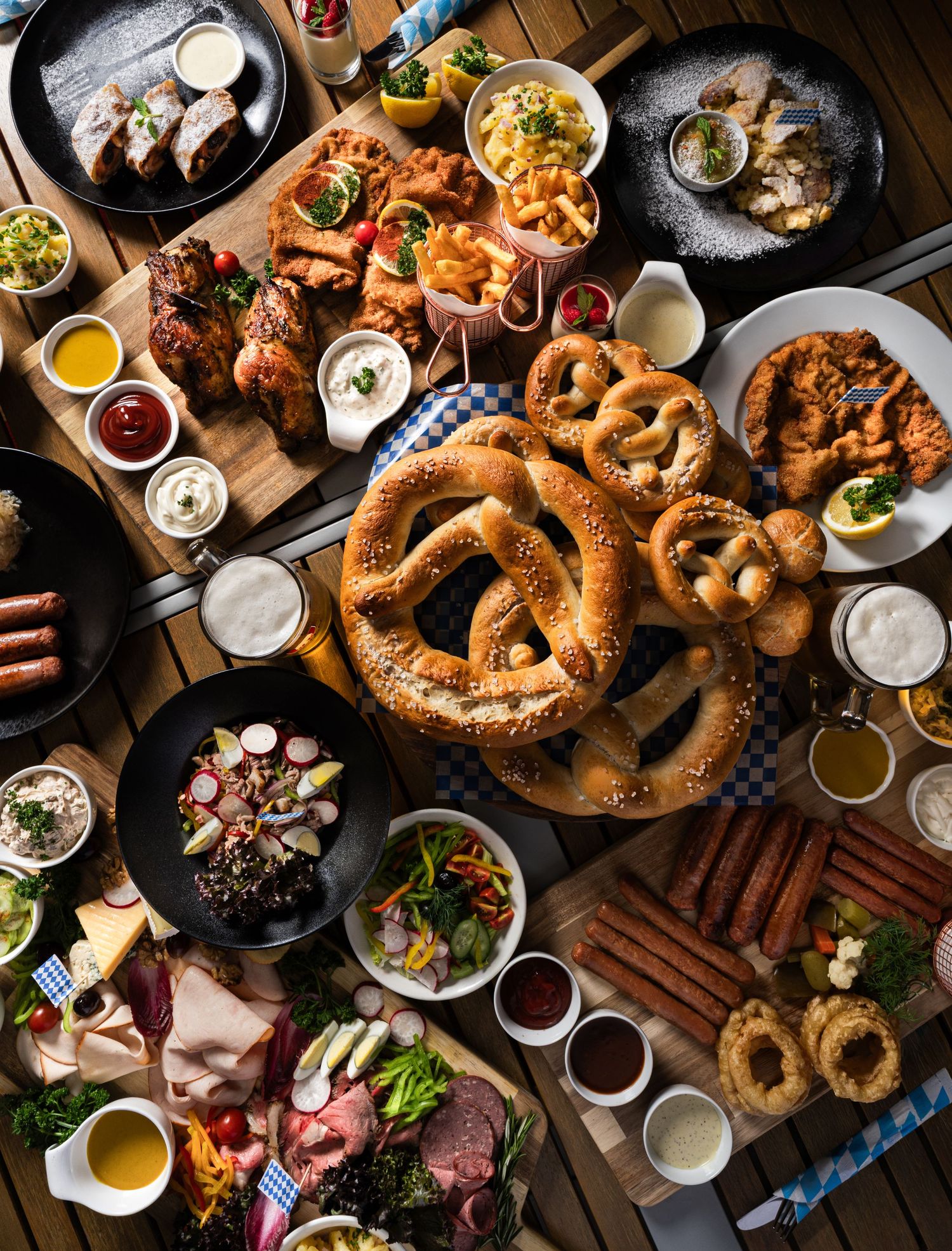 Overhead view of a festive spread with giant pretzels, onion rings, sausages, fried chicken, dips, and charcuterie on a wooden table.