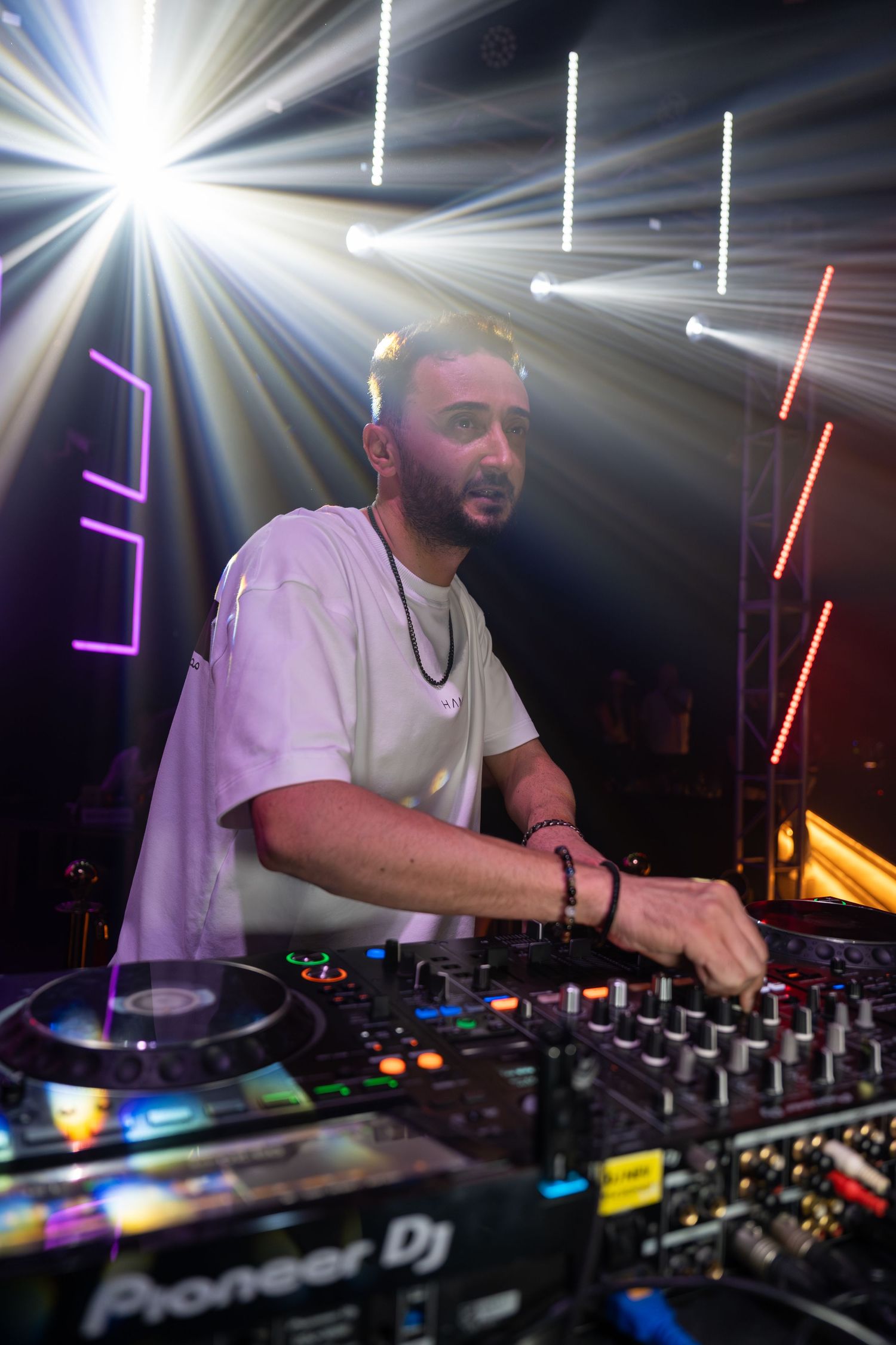 A DJ performs at a nightclub using a Pioneer DJ controller, surrounded by dramatic stage lighting and light beams.