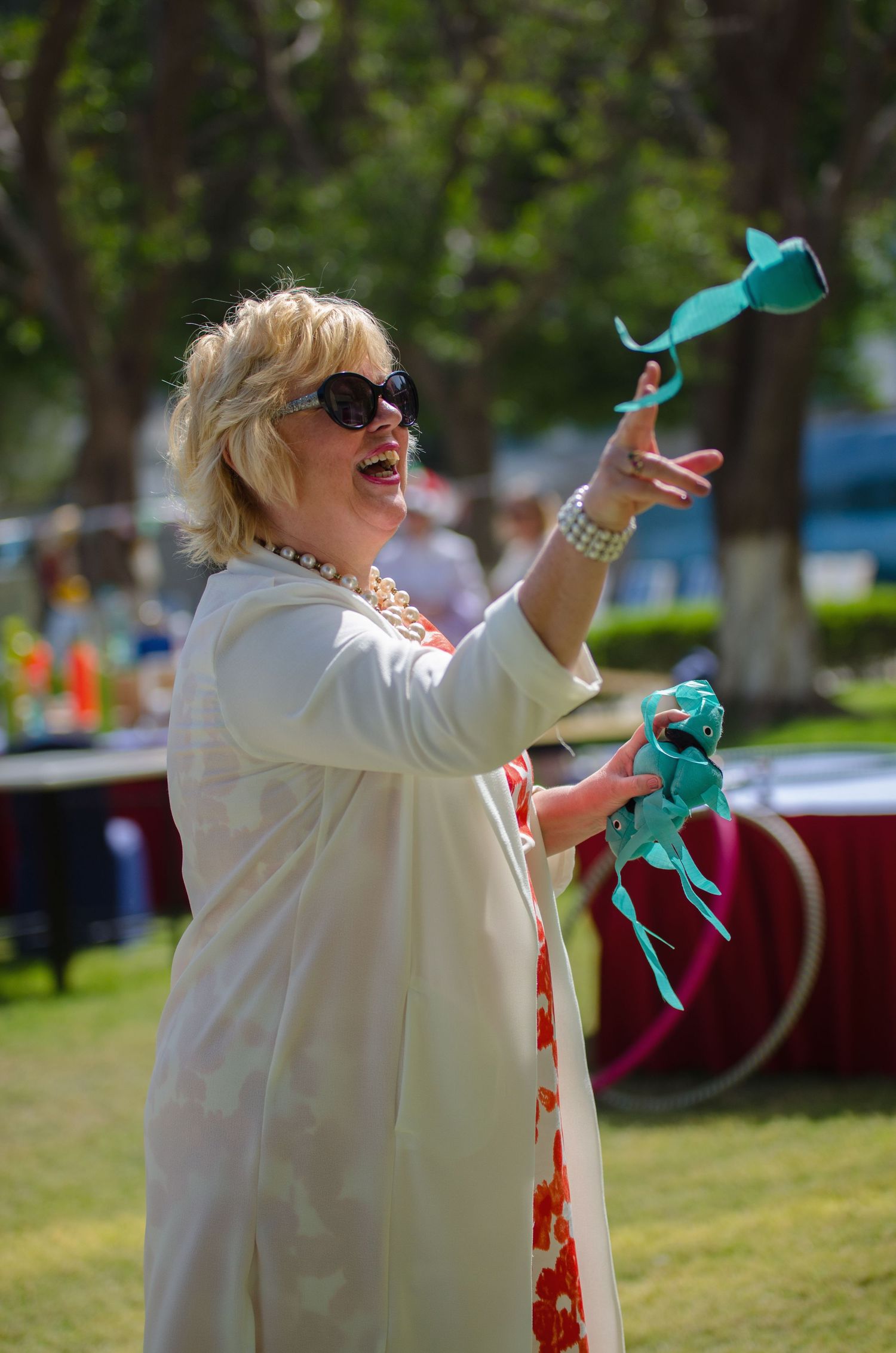 Smiling woman in sunglasses and a white coat tosses teal fabric items outdoors at a sunny park event with tents and trees behind her.