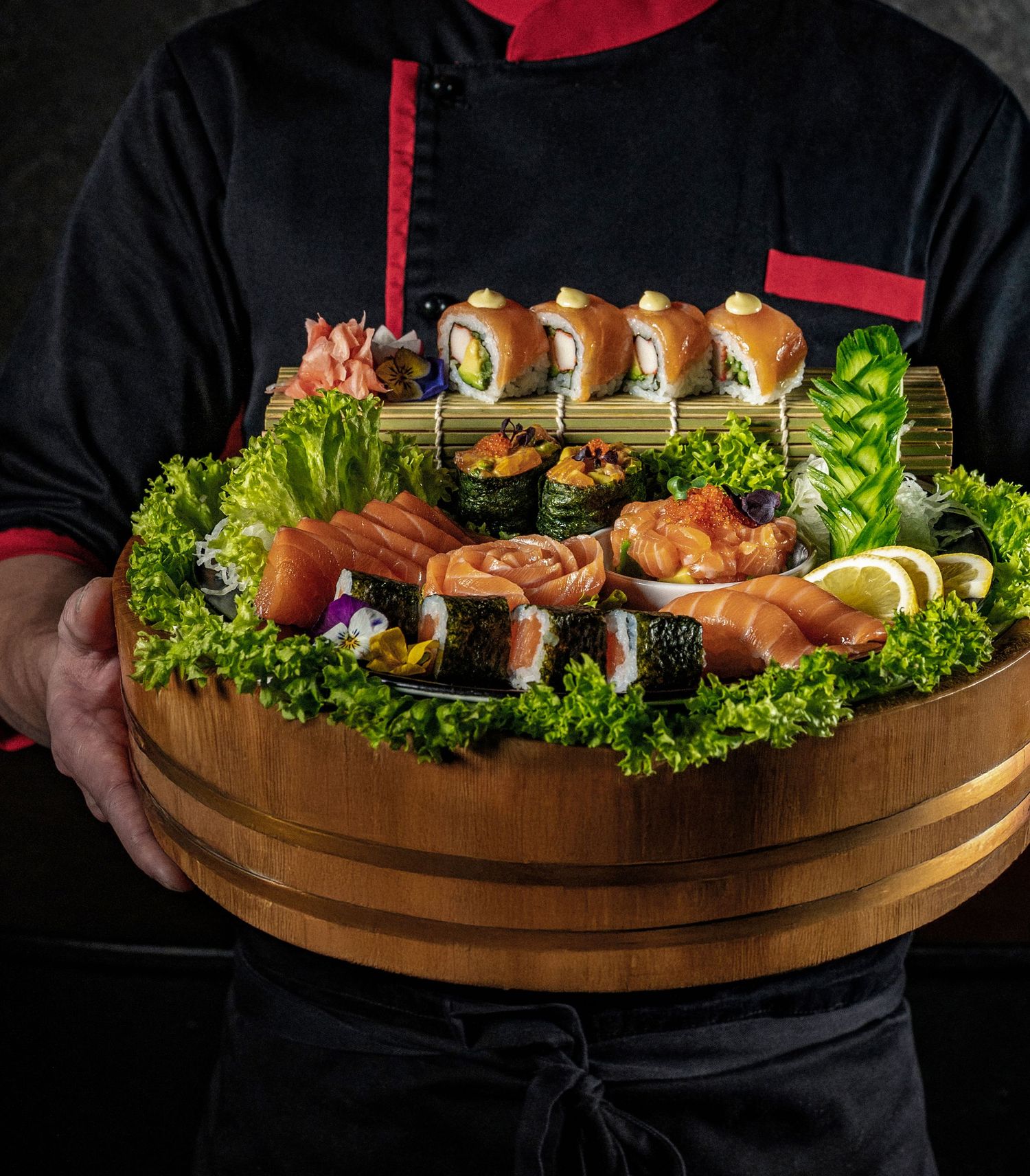 Chef holding a wooden platter filled with assorted sushi rolls, sashimi, and garnishes on fresh green lettuce.