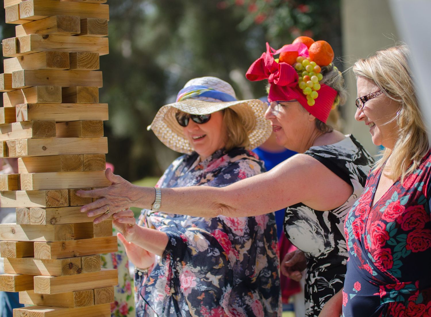 Women in sun hats and floral dresses play a giant outdoor Jenga game, carefully removing a block from a tall wooden tower.