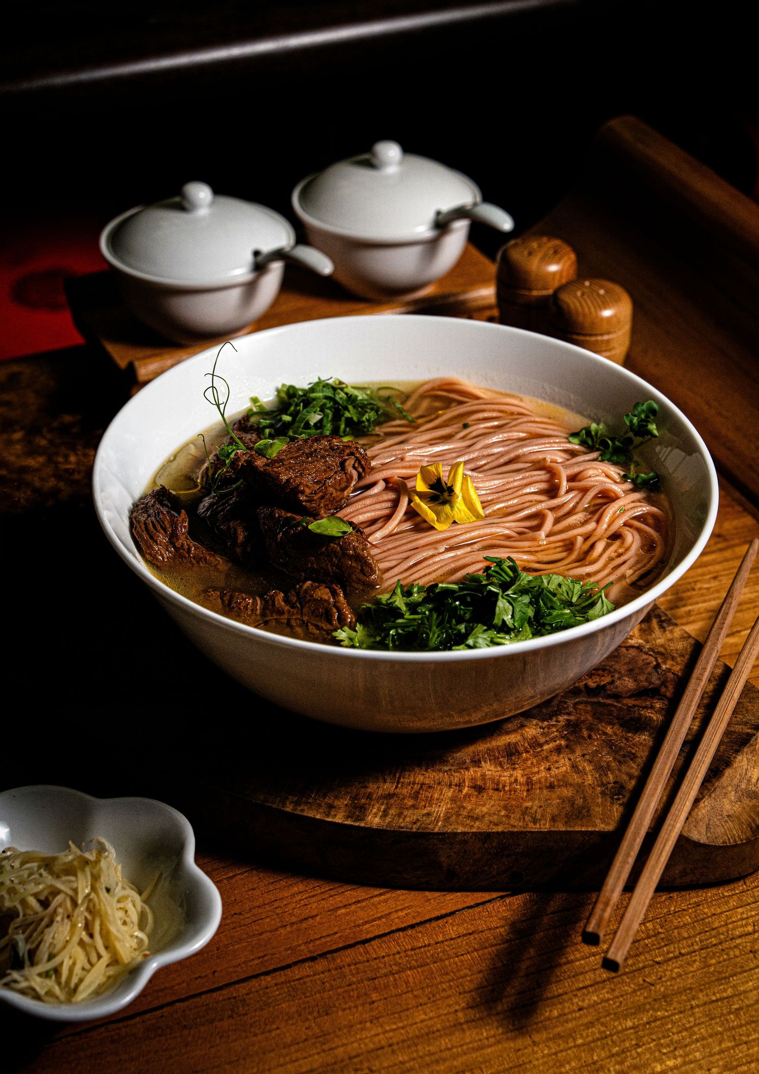 A steaming bowl of Asian noodle soup with braised meat, fresh herbs, and chopsticks on a rustic wooden board.