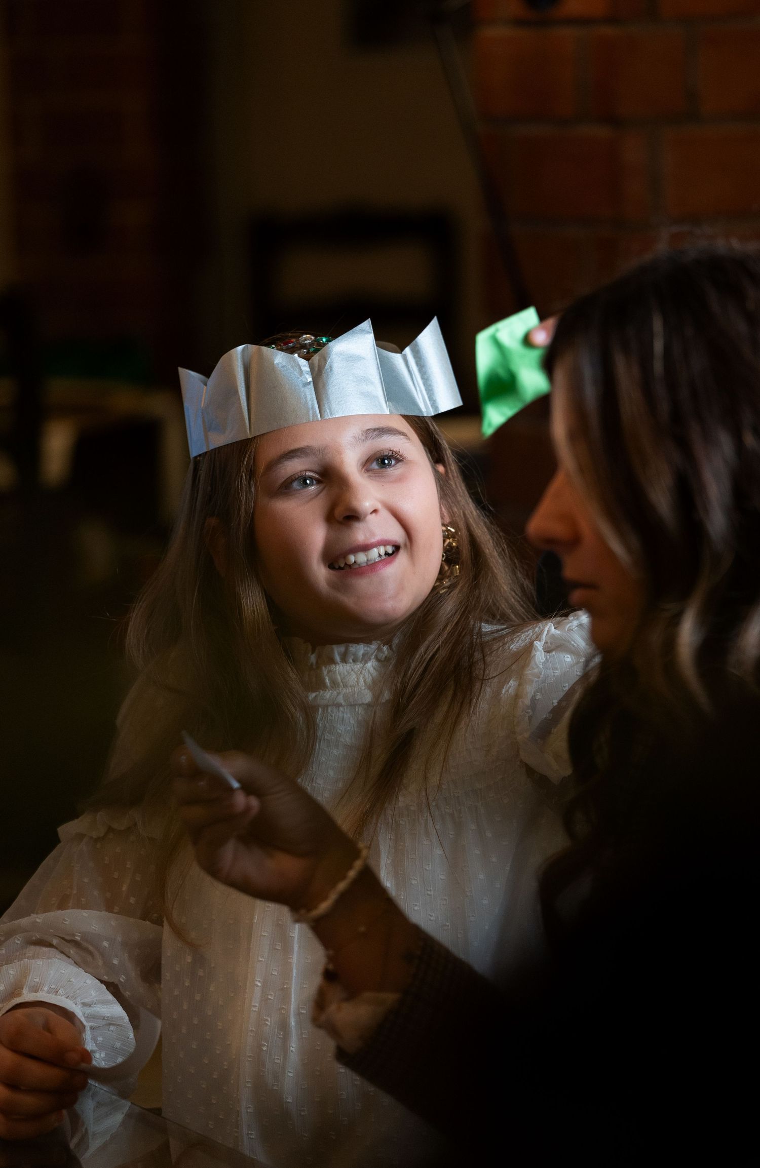 Young girl in white dress and paper crown smiles during First Communion ceremony with woman beside her.