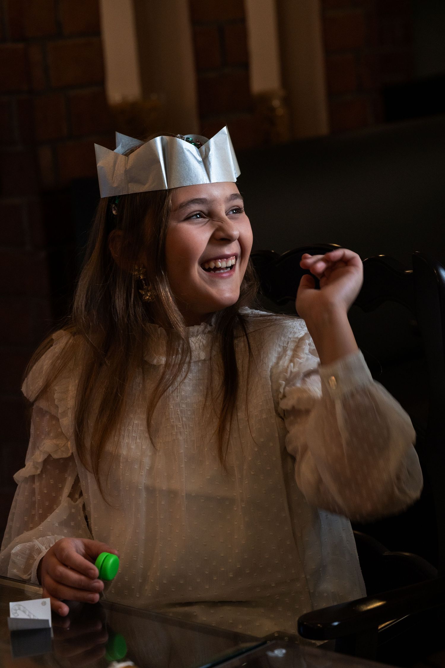 Laughing girl wearing a silver paper crown and white dress, joyfully celebrating at a festive holiday party.