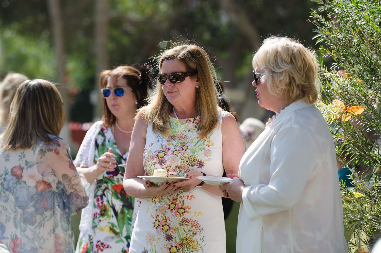 Women in floral dresses and sunglasses chat outdoors at a sunny garden party, one holding a plate of food among green trees.