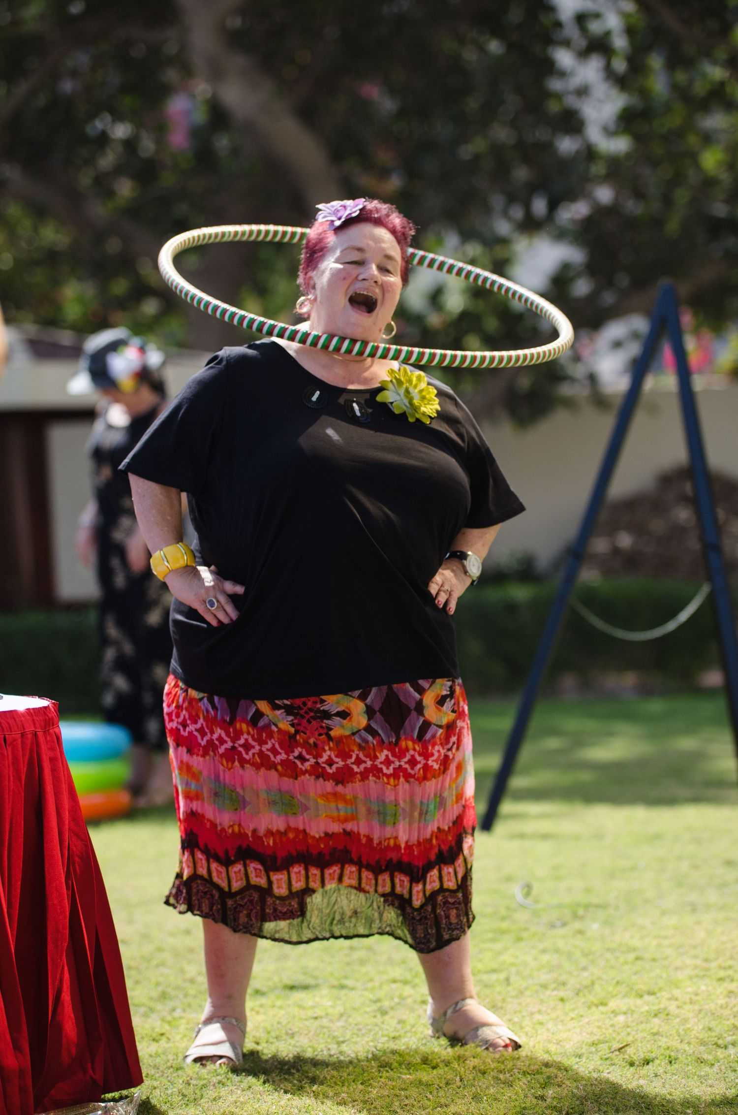 Woman in a black top and colorful skirt laughs while spinning a hula hoop around her neck on a sunny backyard lawn.