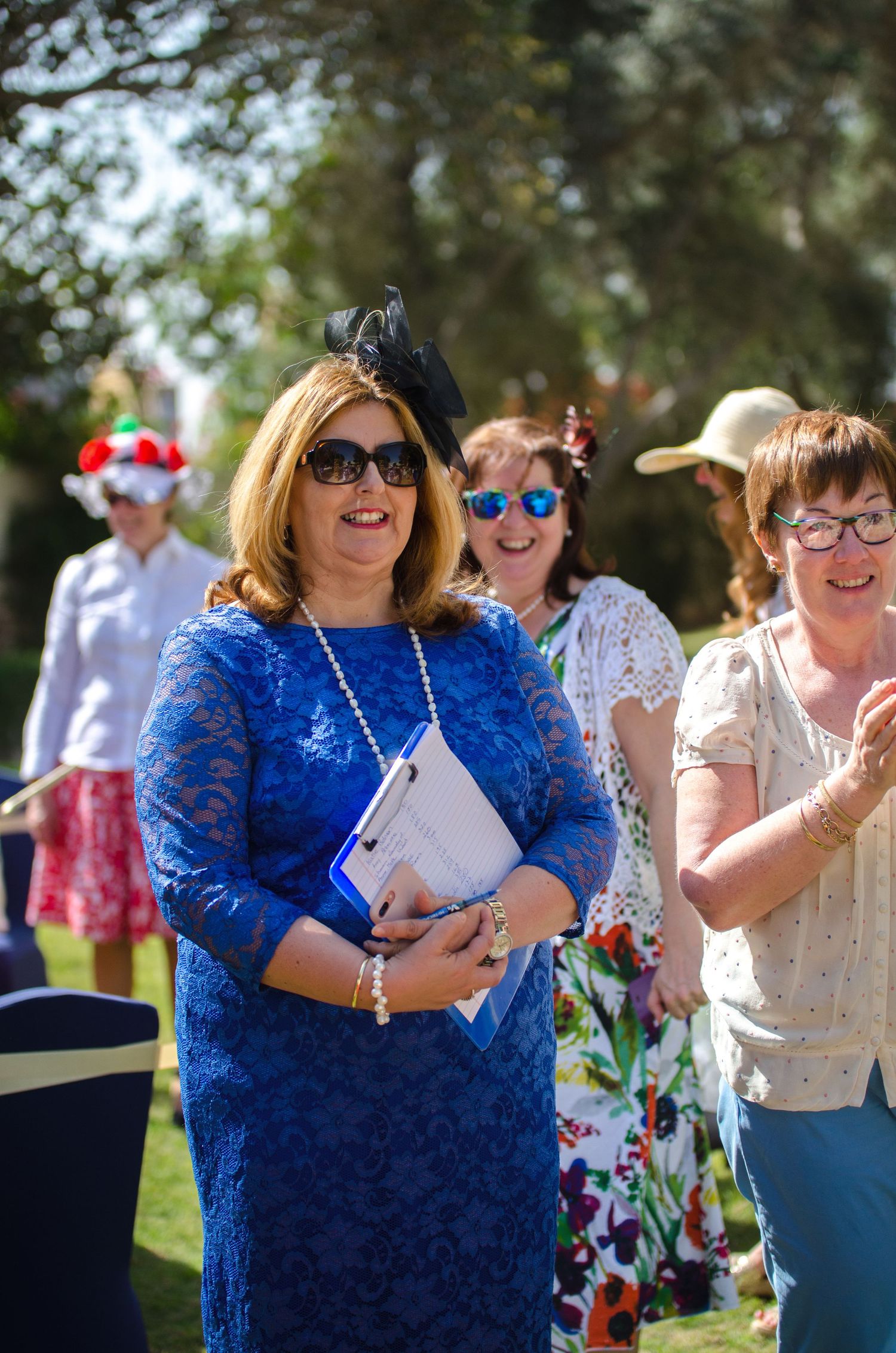 Smiling woman in a blue lace dress, sunglasses and black fascinator holds papers at an outdoor garden event with other guests behind her.