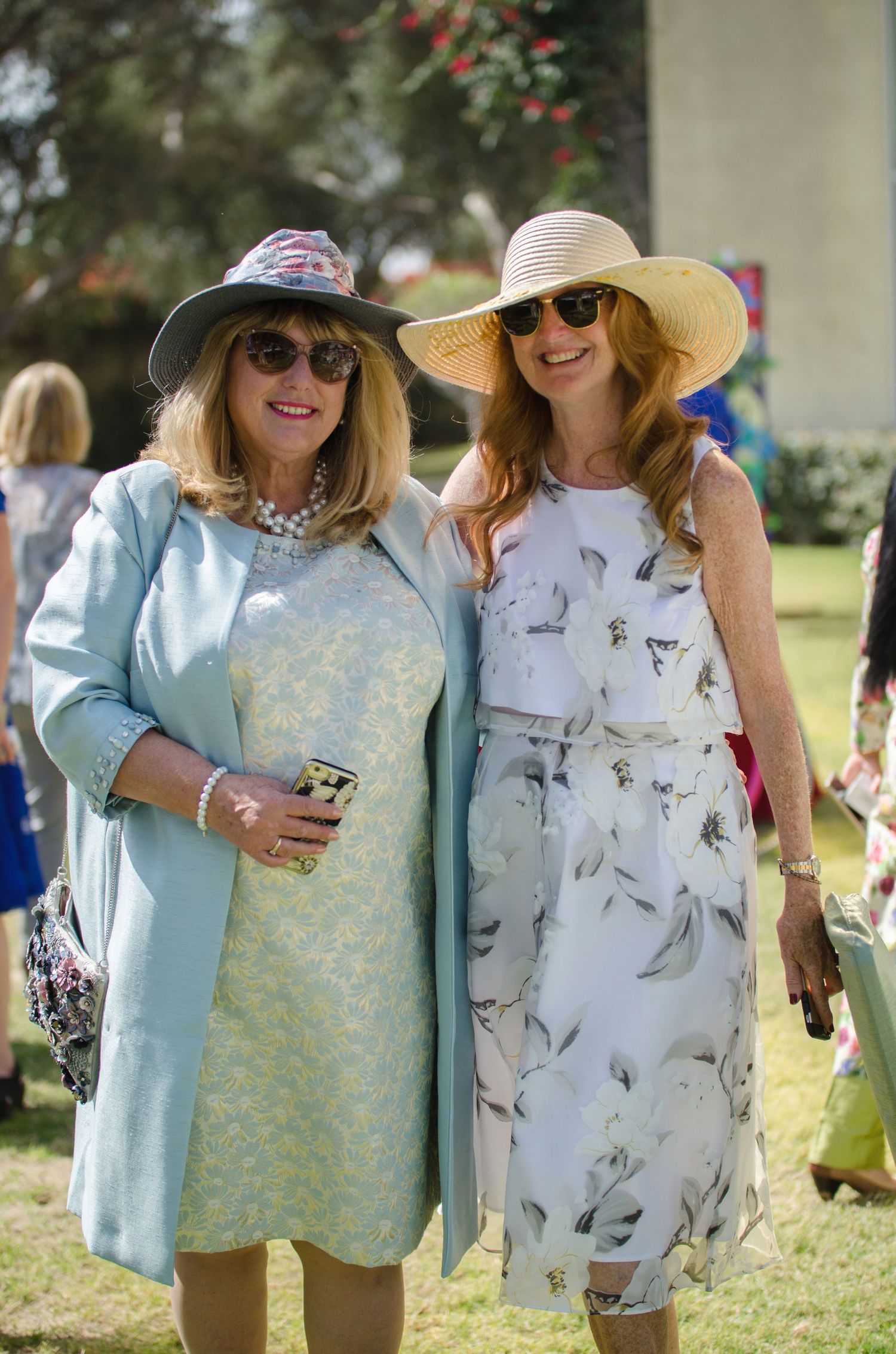 Two smiling women in sunglasses and stylish hats pose arm in arm in floral dresses at an outdoor garden event on a sunny day.
