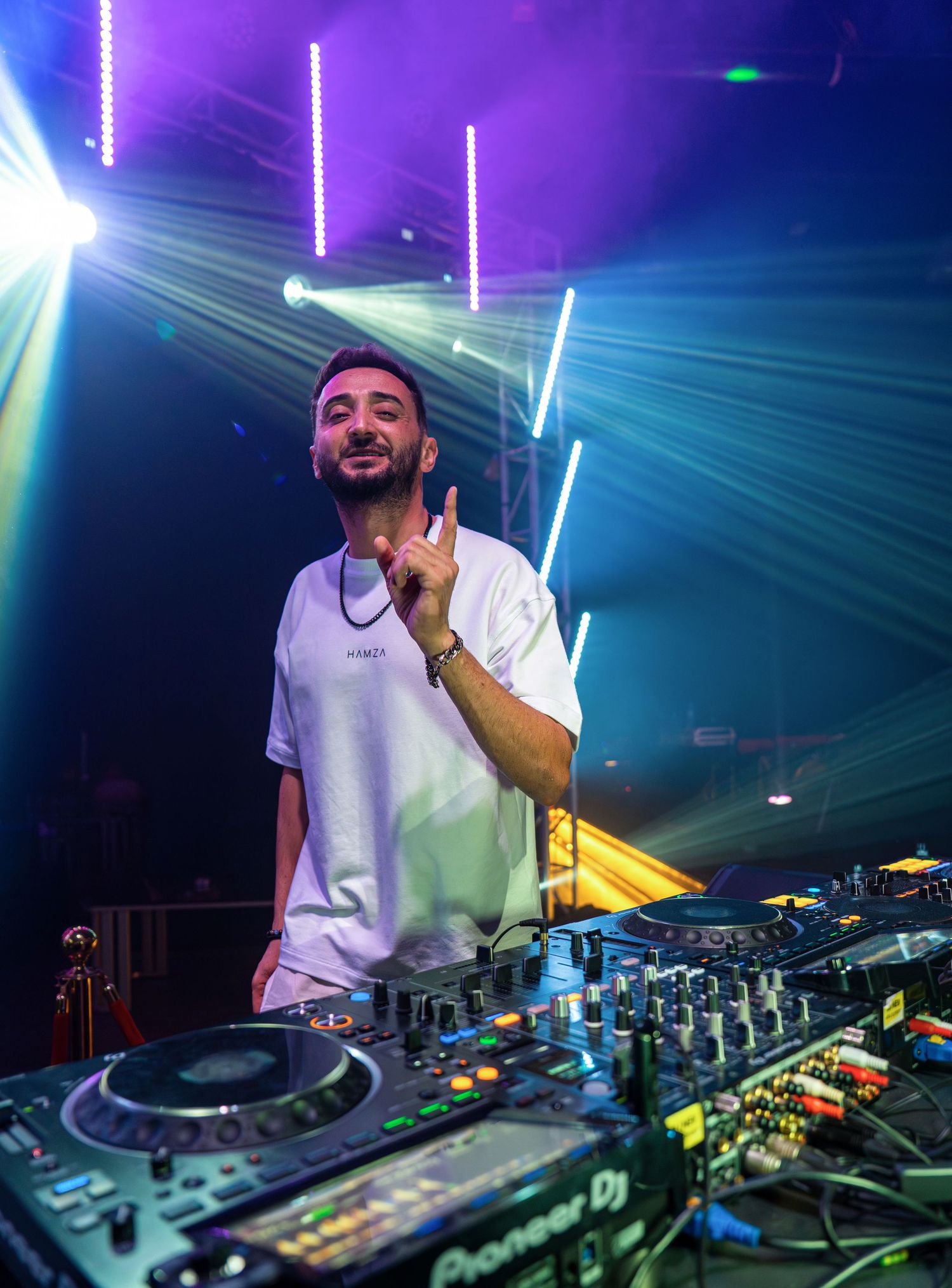 Male DJ gesturing with one finger raised behind Pioneer DJ decks at a nightclub event.