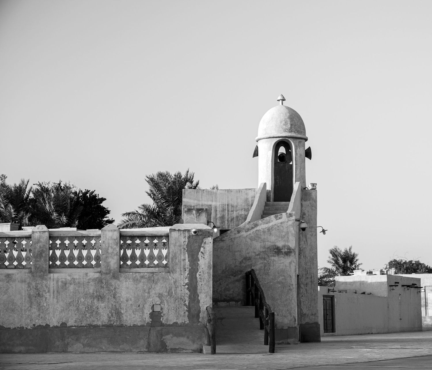 Black and white photo of an ancient Middle Eastern building with a domed minaret, decorative wall, and stairs.