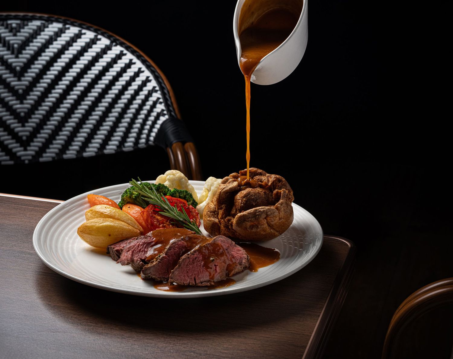 Gravy pouring from a white jug over a roast beef dinner with Yorkshire pudding and roasted vegetables on a white plate.