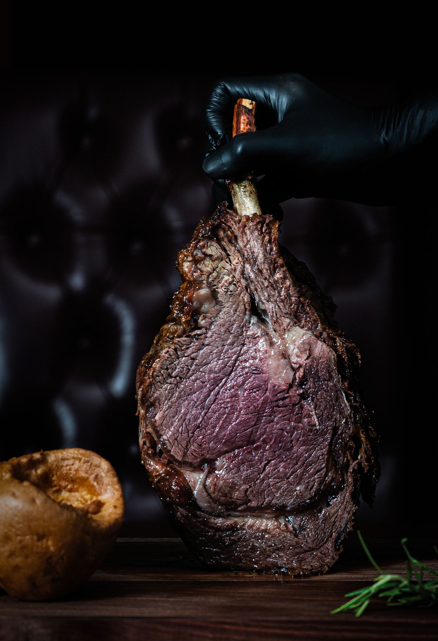 A gloved hand holds a perfectly roasted bone-in prime rib on a wooden board, with a Yorkshire pudding and rosemary.