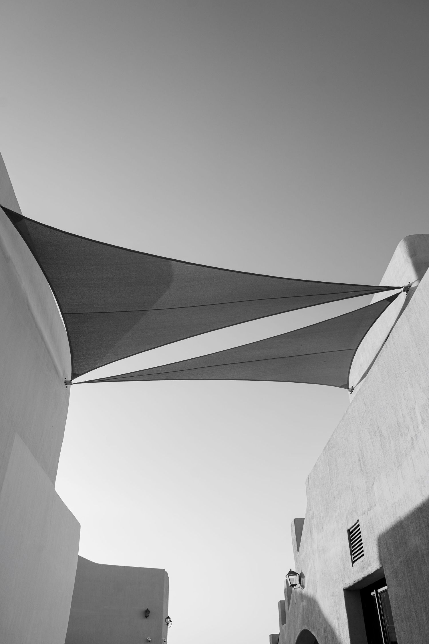 Black and white photo of a triangular shade sail stretched between white adobe buildings against a clear sky.