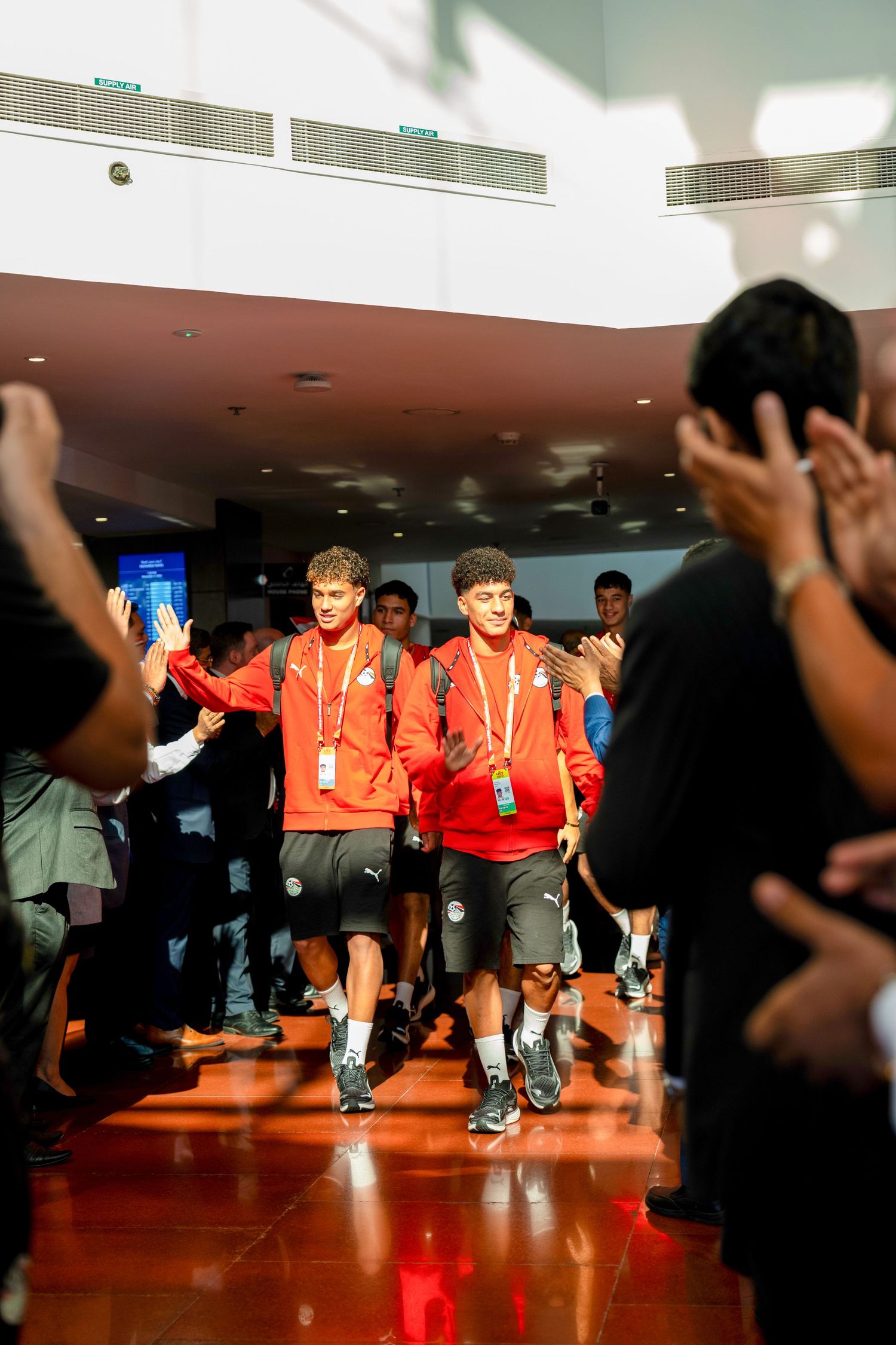 Young athletes in red uniforms walking through a crowd of cheering fans indoors.