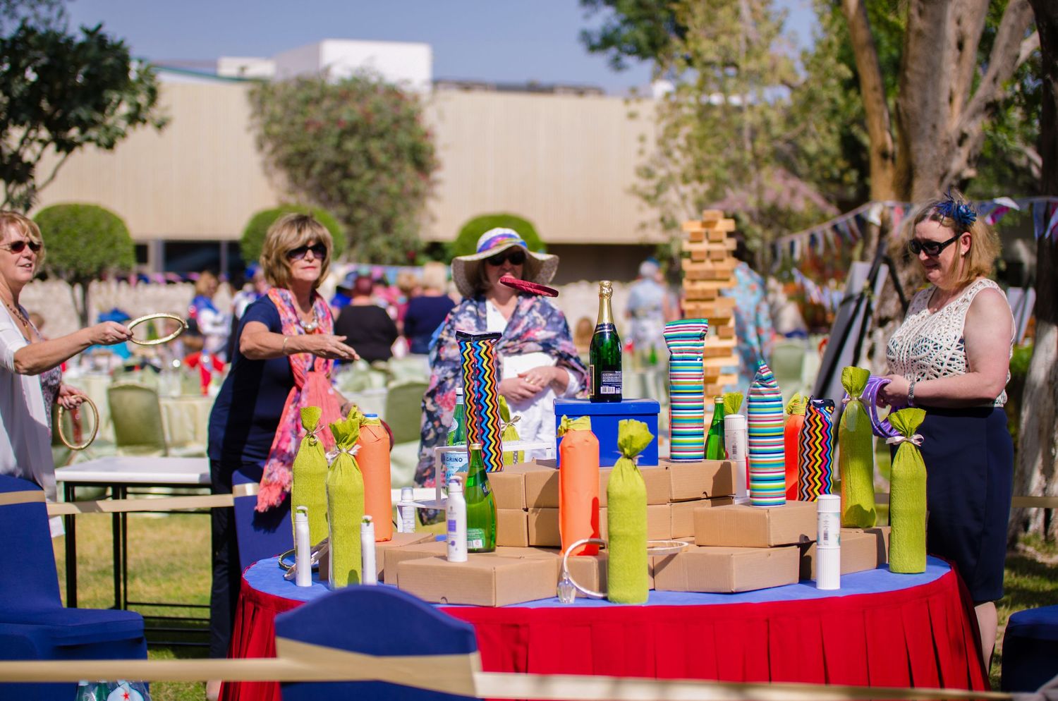 Women in summer dresses browsing colorful outdoor game tables with Jenga and festive decorations at a garden party.