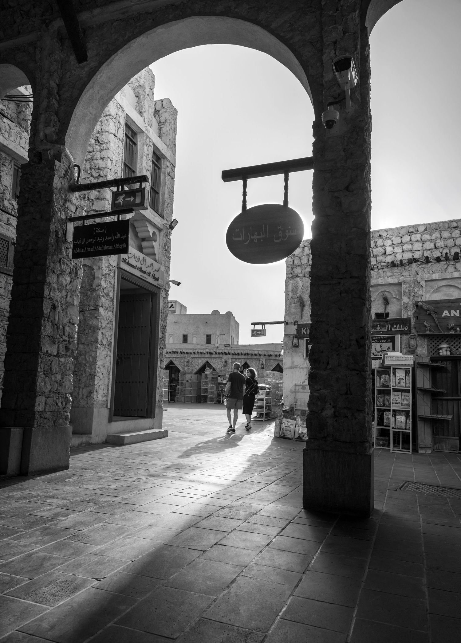 Black and white photo of a stone archway leading to a Middle Eastern marketplace with two people walking.