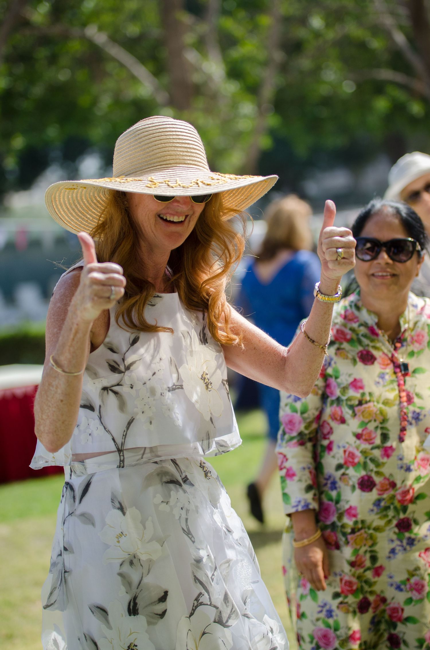Smiling woman in a straw sun hat and floral dress gives two thumbs up at an outdoor garden party while guests mingle nearby.