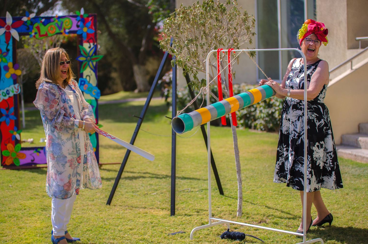 Two women play a colorful striped percussion instrument on a sunny lawn, one in a bright floral wig and dress, near a vibrant art display.