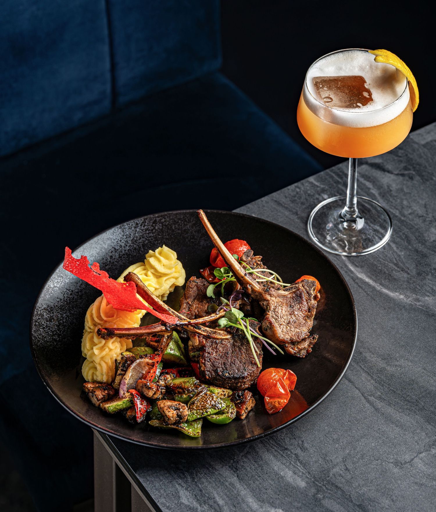 Elegant lamb chops with roasted vegetables and mashed potatoes on black plate beside a frothy cocktail in coupe glass.