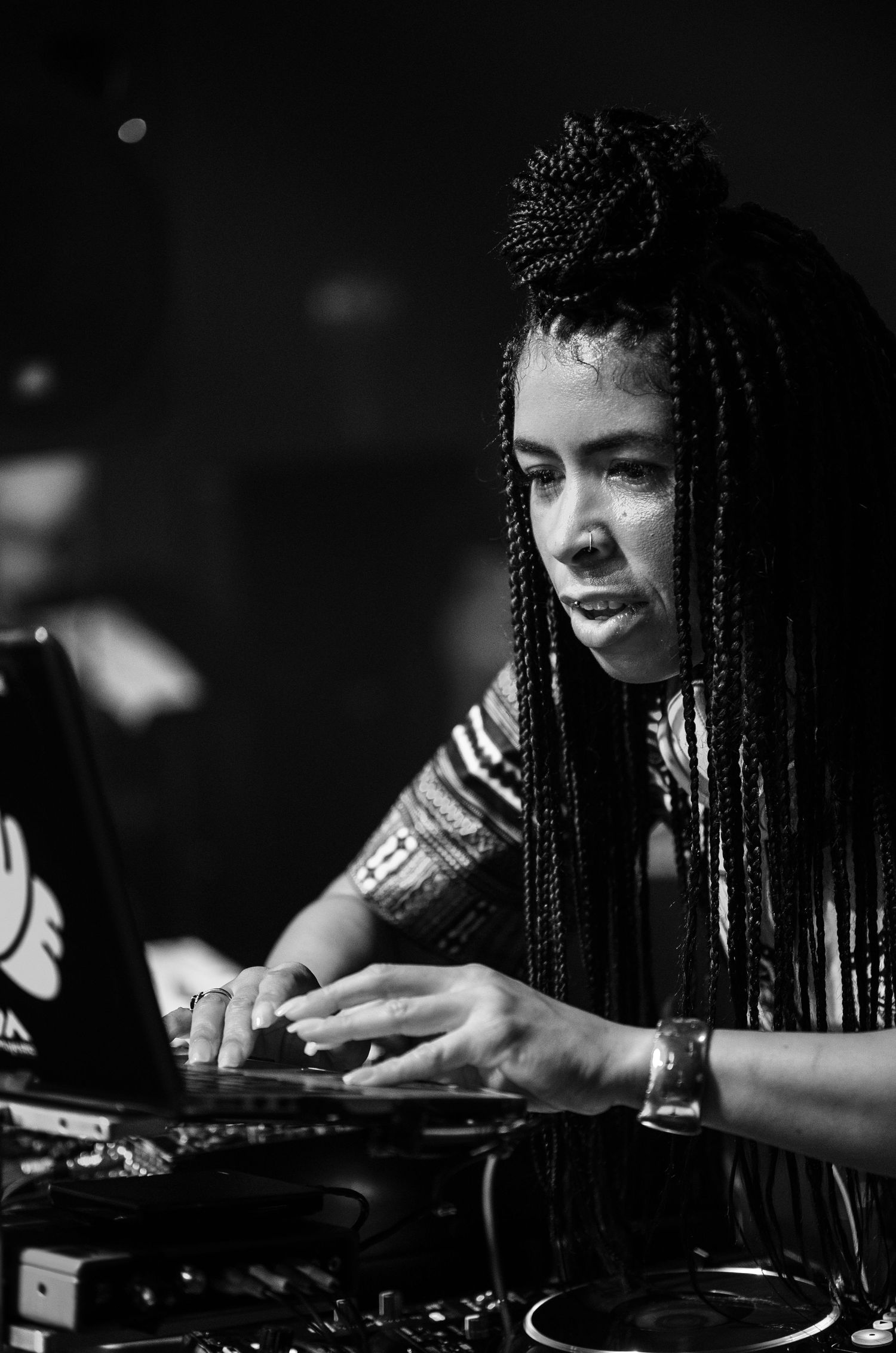 Black and white photo of a female DJ with long braids mixing music on turntables at a live event.