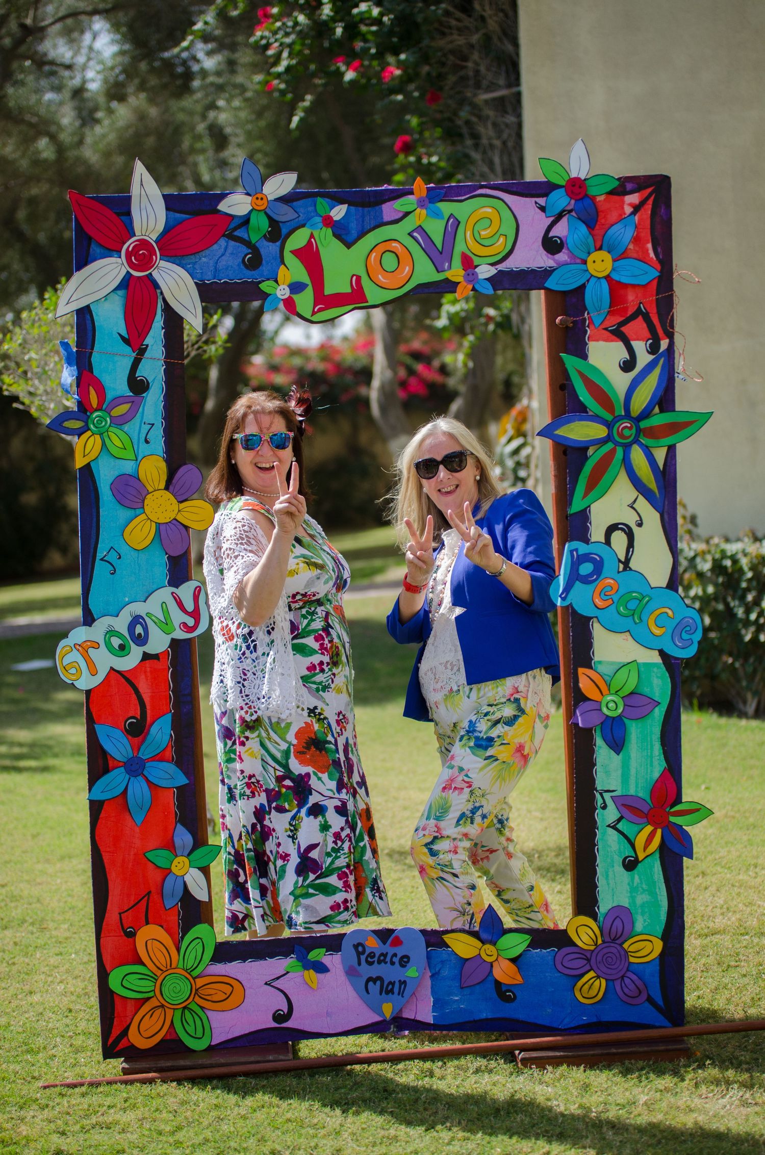 Two smiling women in floral outfits flash peace signs inside a colorful hand-painted frame with flowers, love, groovy and peace lettering.