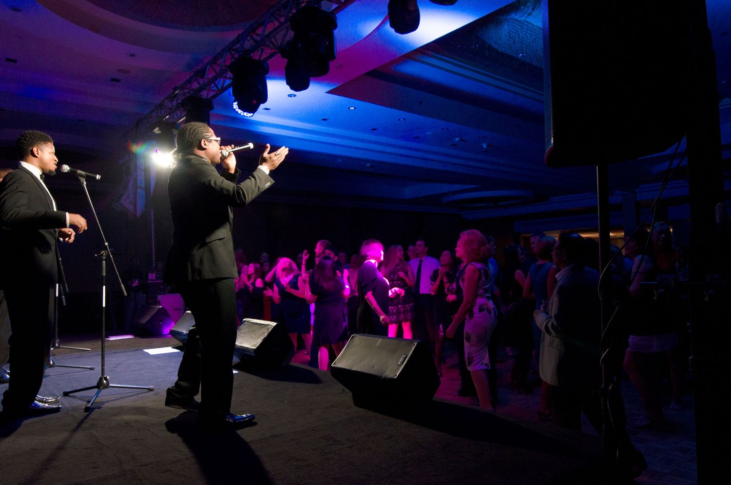 Male performers in black suits singing on a blue-lit stage to an engaged crowd at a formal gala.