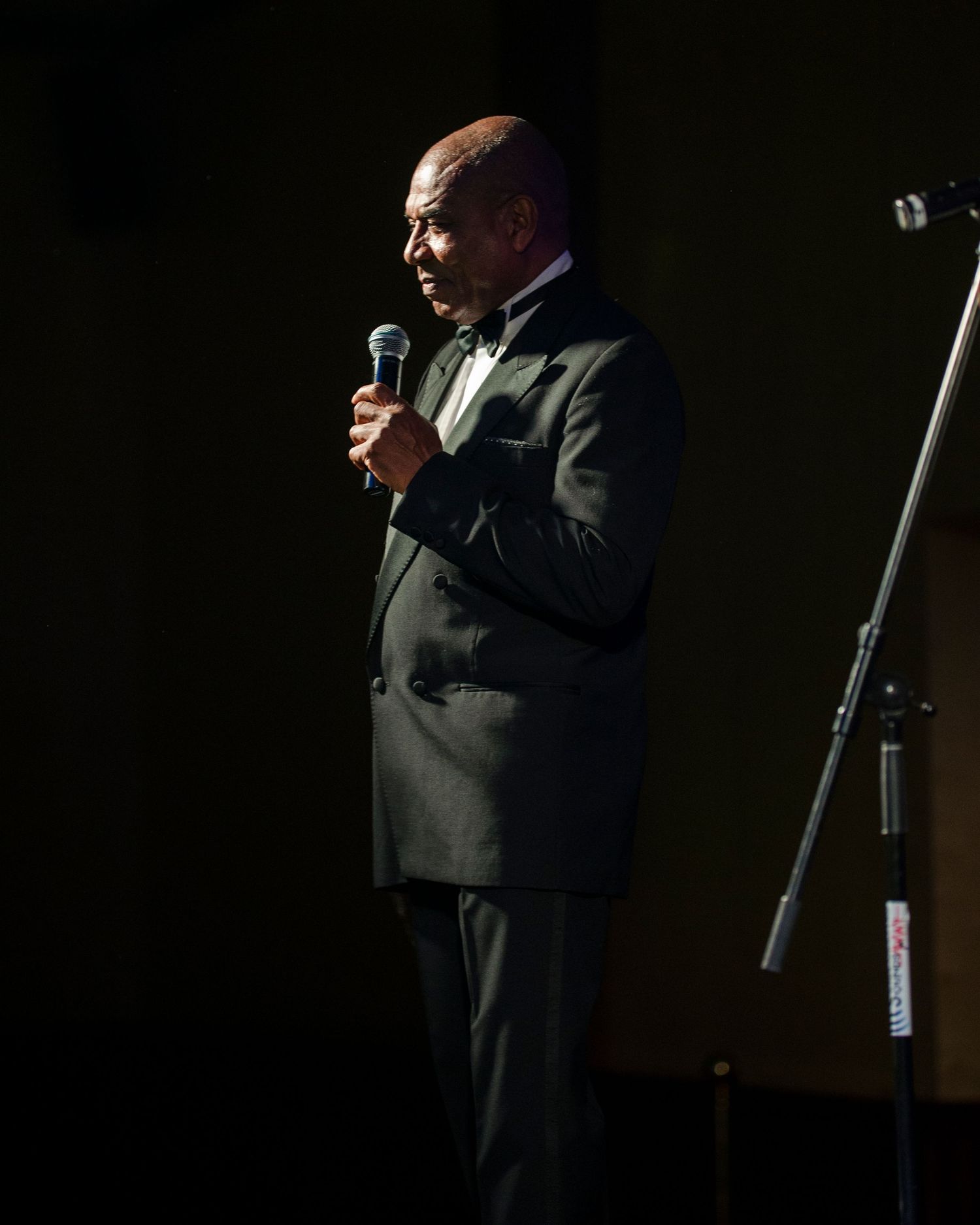 Bald man in black tuxedo speaking into a microphone on a dimly lit stage at a formal event.