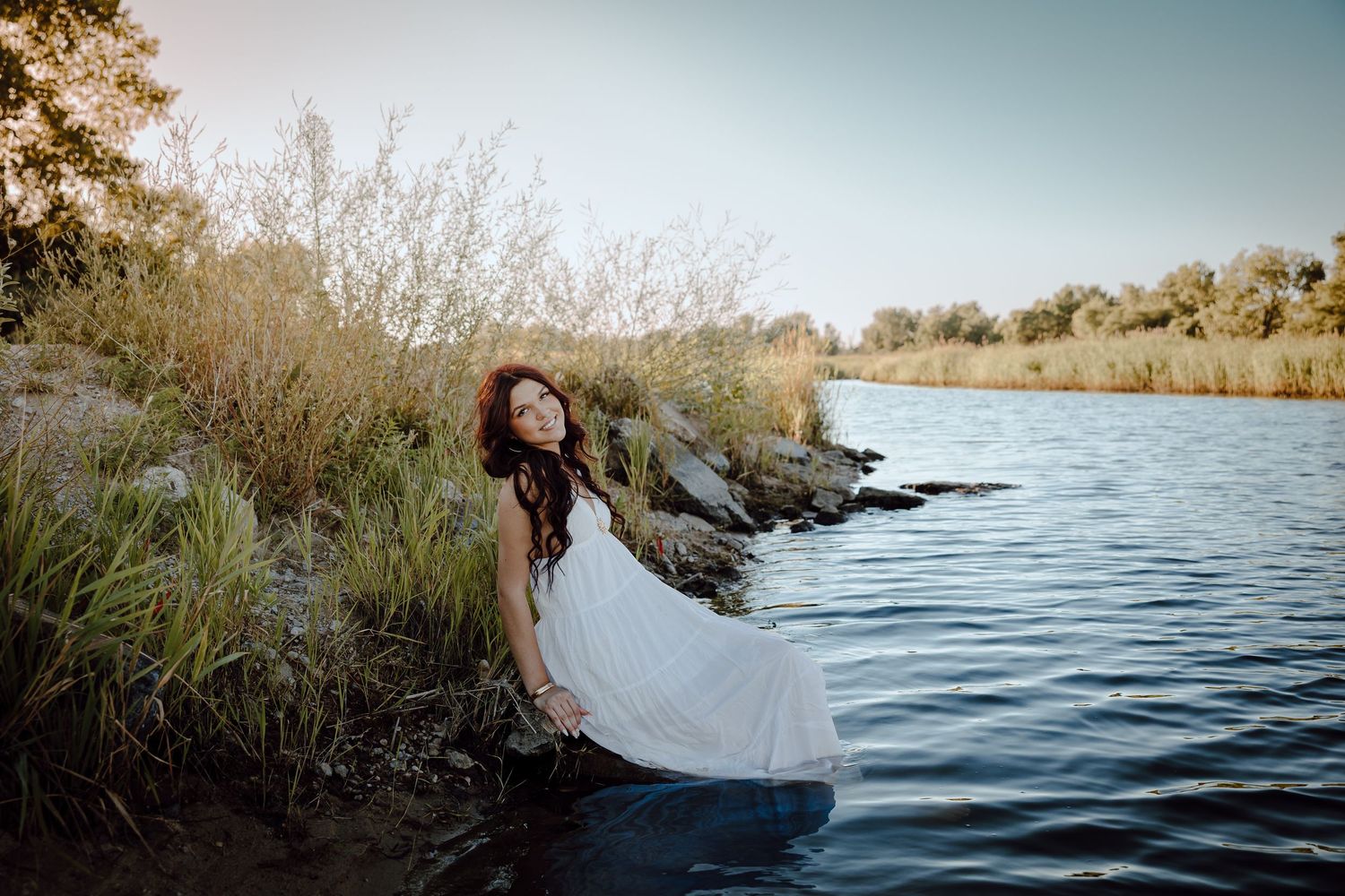 western Nebraska senior session a girl in a white dress sits at the water's edge surrounded by lush greenery and rocks at golden hour.