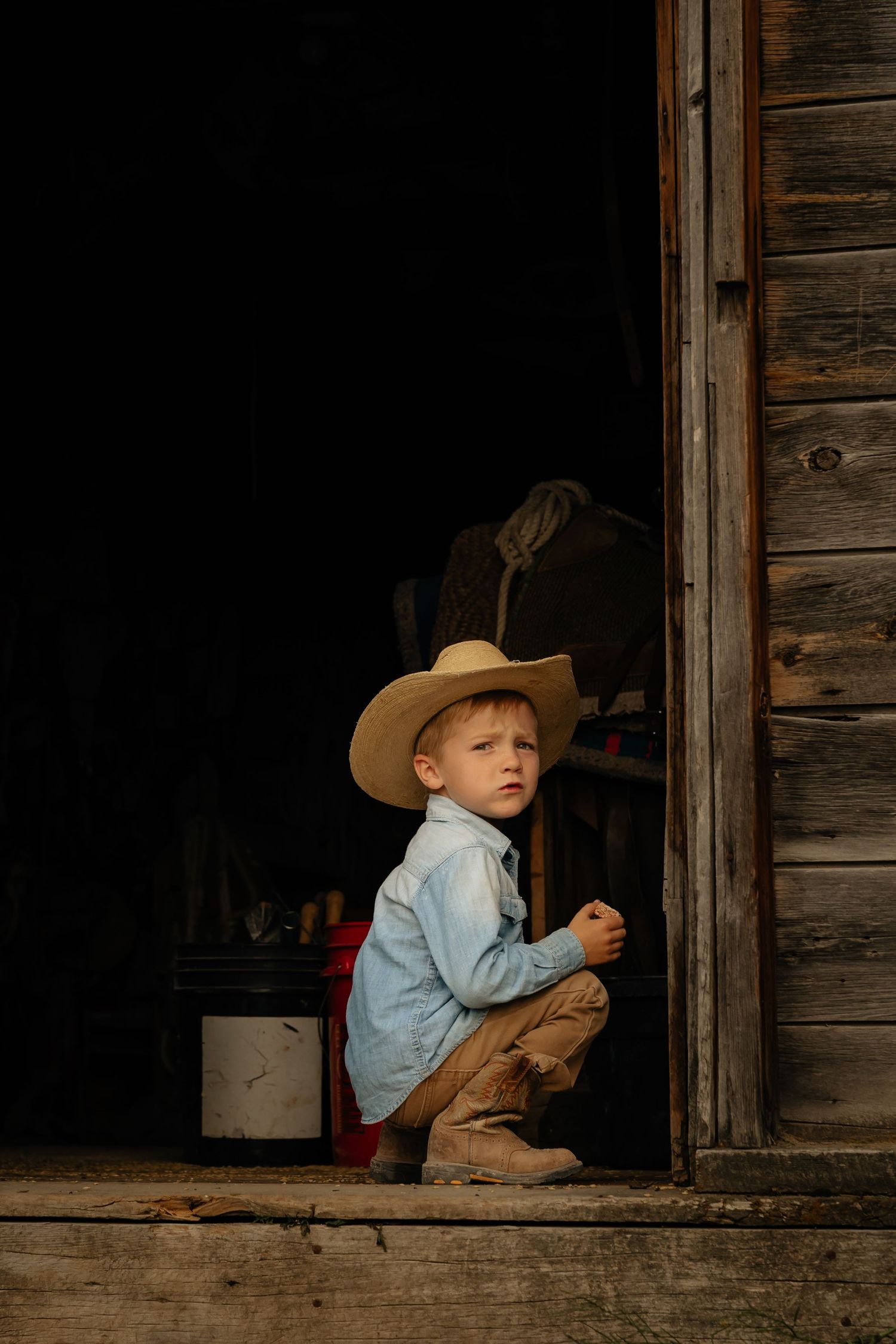 Western Nebraska Family Sessions boy in cowboy hat and blue shirt crouching in rustic barn doorway, bathed in warm golden light.