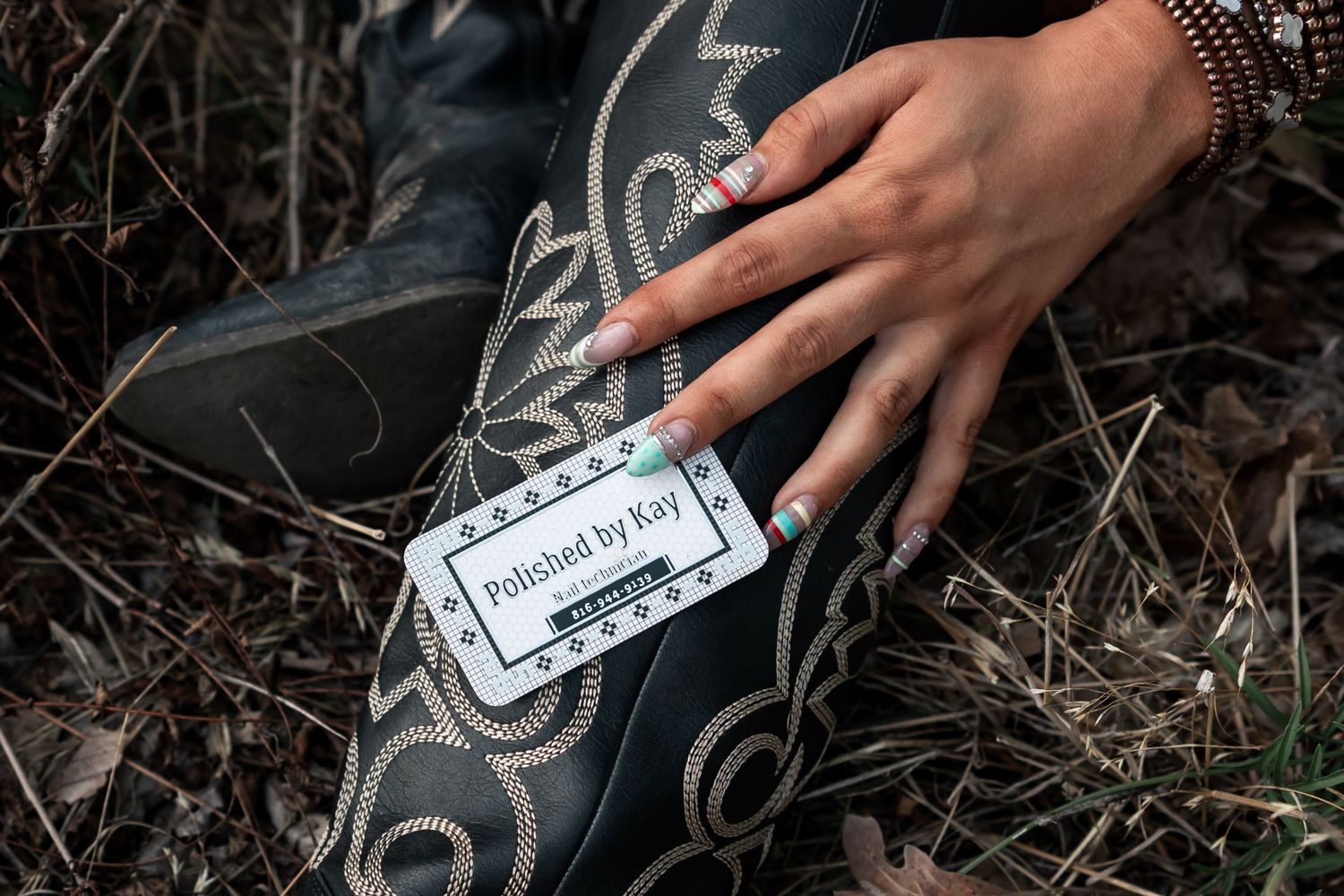 Western Nebraska Brand Session hand with decorated nails holding a 'Polished by Kay' business card against black cowboy boots outdoors.
