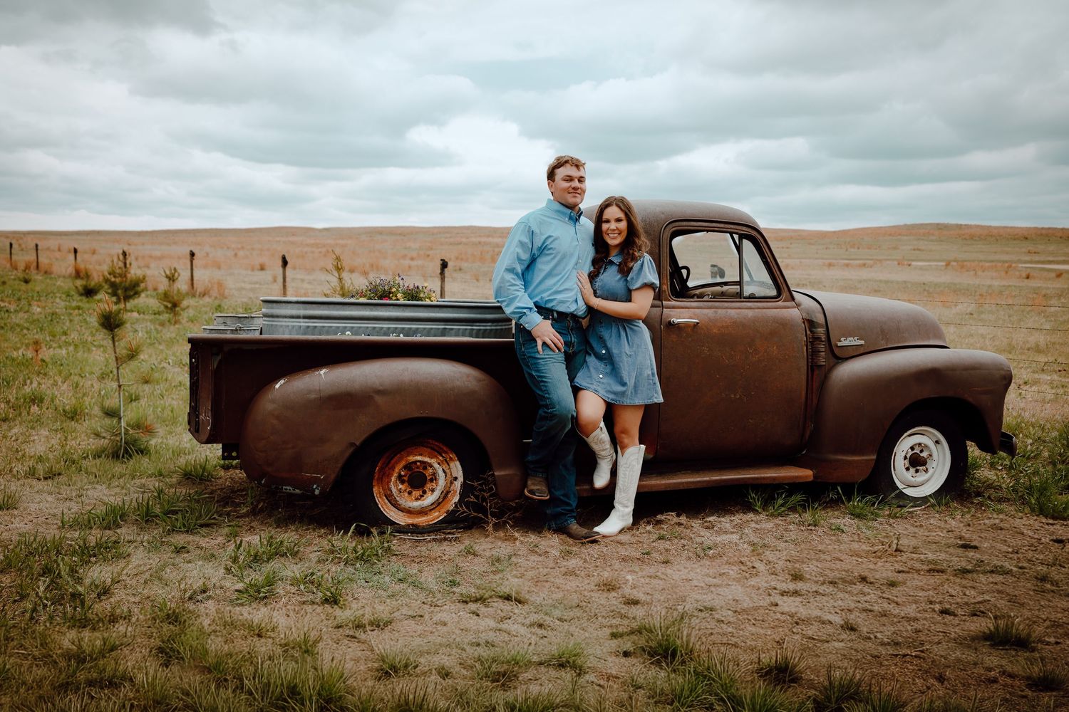 Nebraska Sandhills Engagement Session Couple posing by a rustic vintage brown pickup truck in an open field under a cloudy sky.
