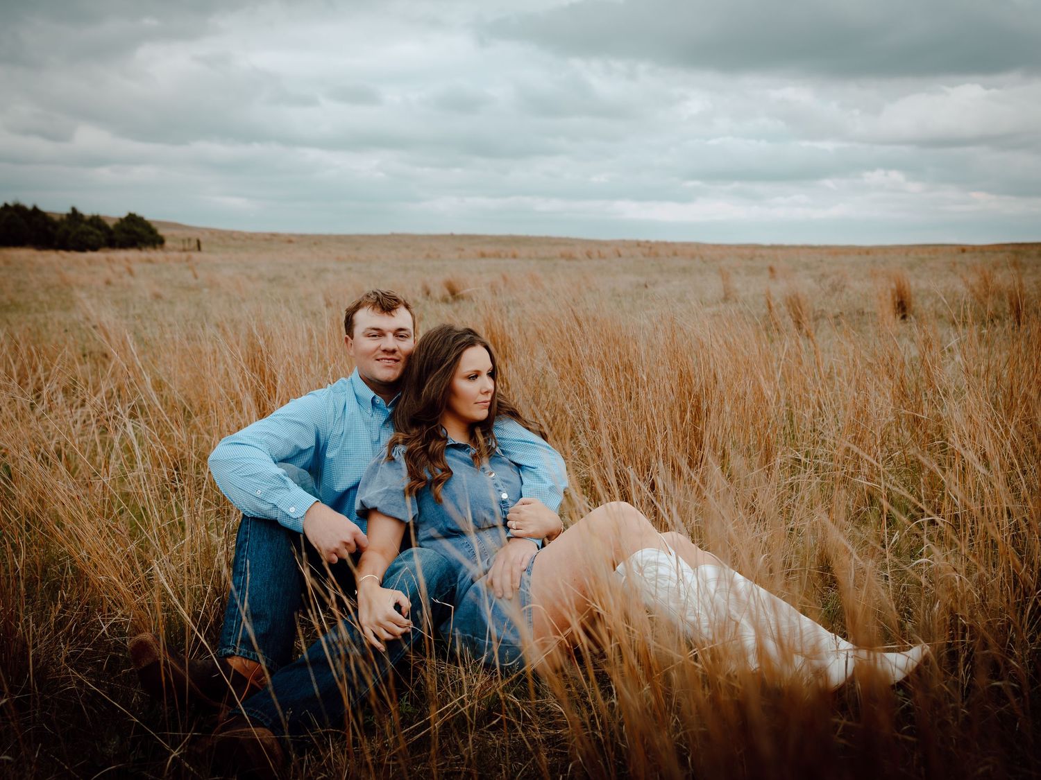 Nebraska Sandhills engagement session couple sitting together in a golden field under cloudy skies, wearing denim outfits