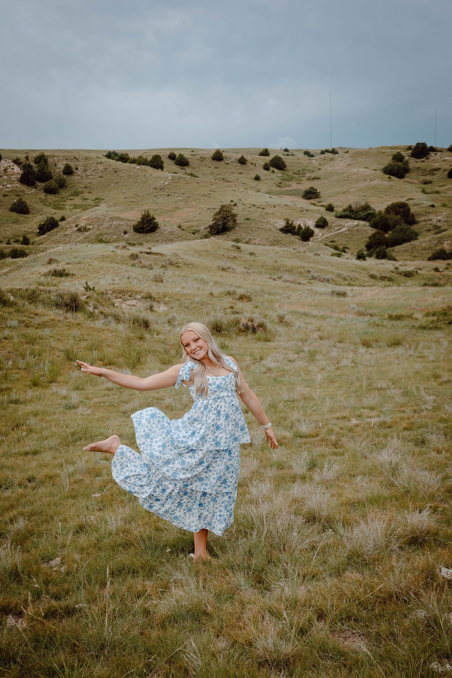 Oshkosh Nebraska senior session girl in floral blue dress dancing barefoot in a grassy meadow with rolling hills under cloudy skies.