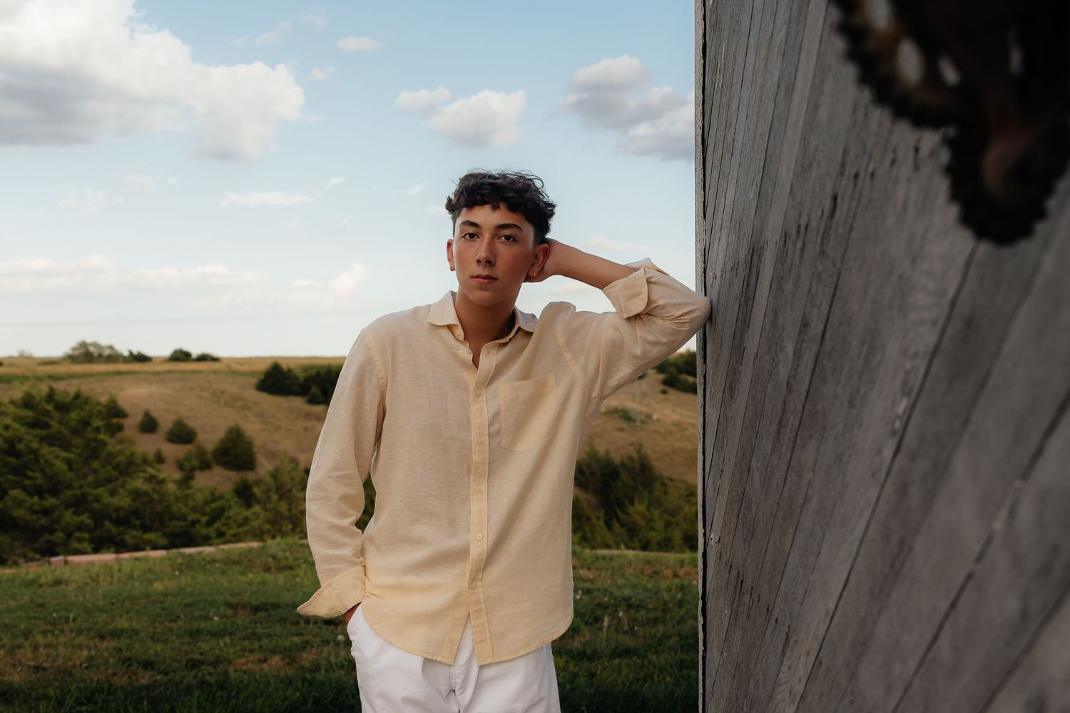 North Platte Nebraska Senior Session young man in yellow shirt leaning against rustic wood wall with green countryside landscape behind him.