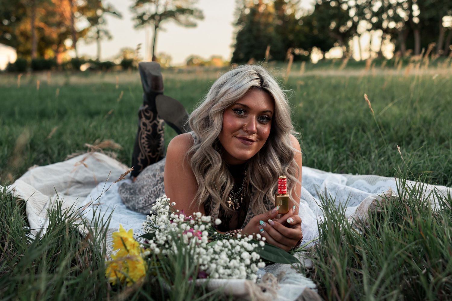 Western Nebraska Brand Session woman lying on a white blanket in a green field, holding a small bottle, surrounded by wildflowers at sunset.