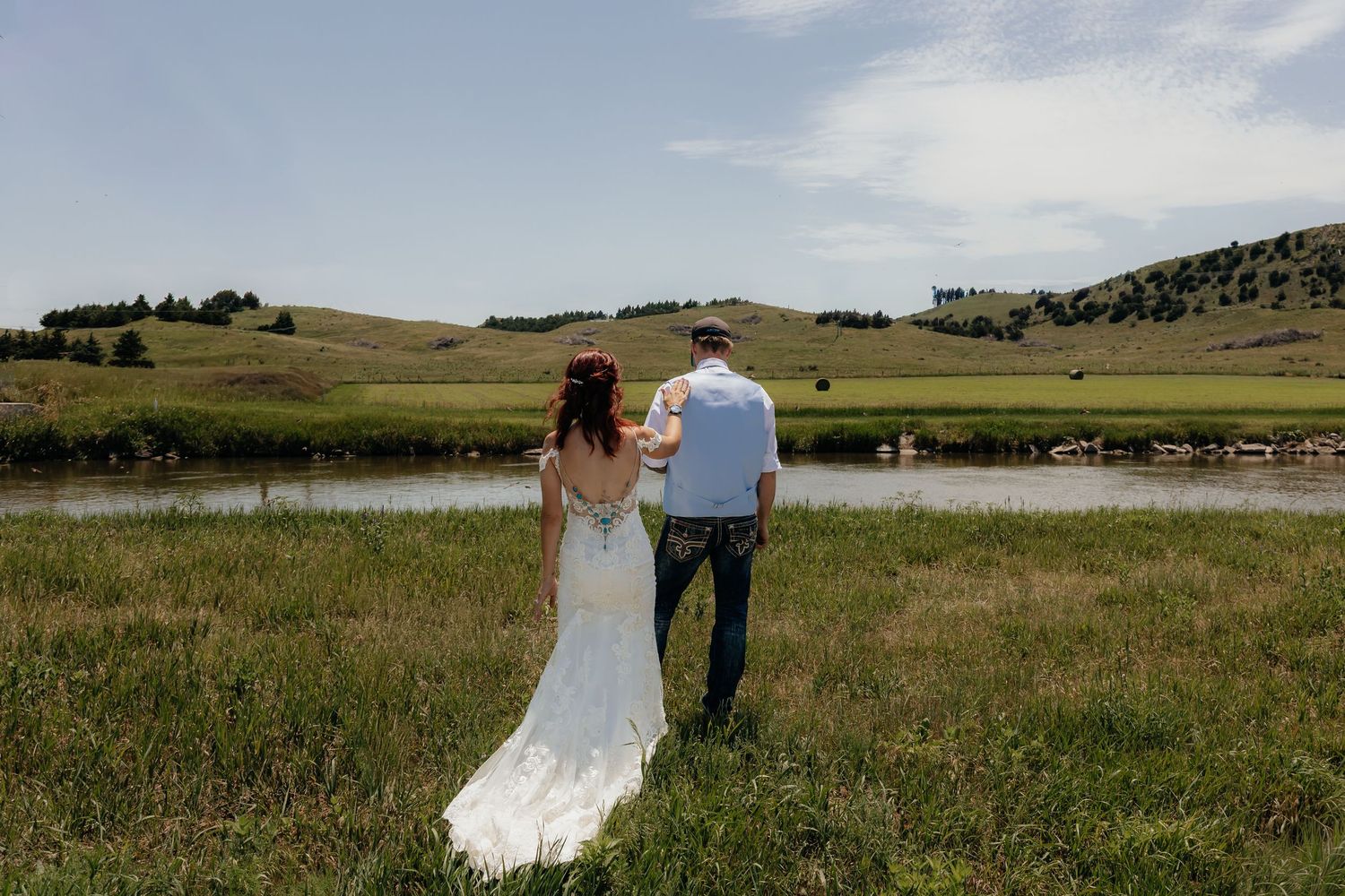 Western Nebraska Wedding Bride and groom standing hand in hand facing a river surrounded by lush green hills